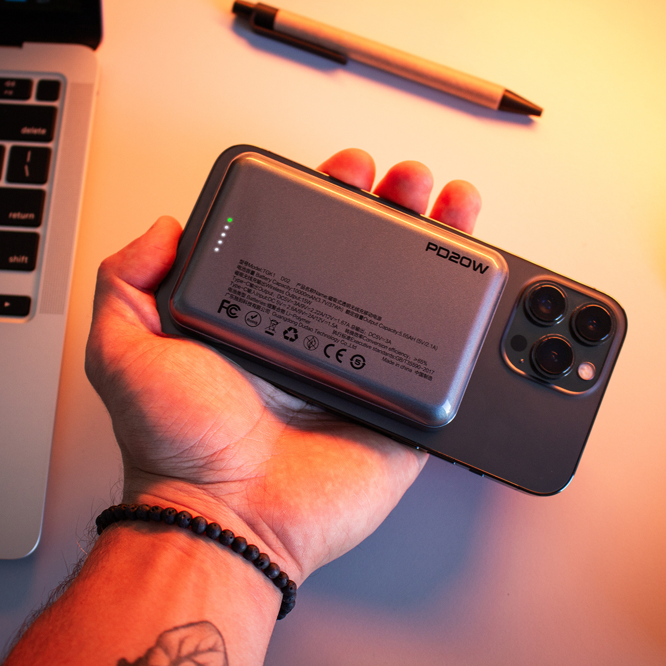 Dłoń trzymająca telefon z dołączonym power bankiem. Laptop i długopis w tle.