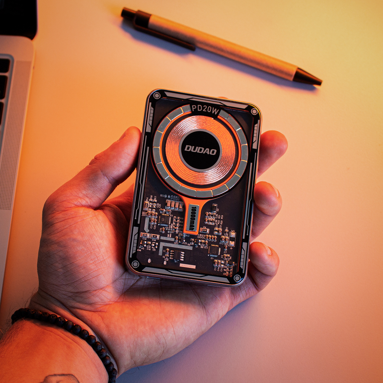 Dłoń trzymająca power bank z przezroczystym przodem. Pióro na stole.