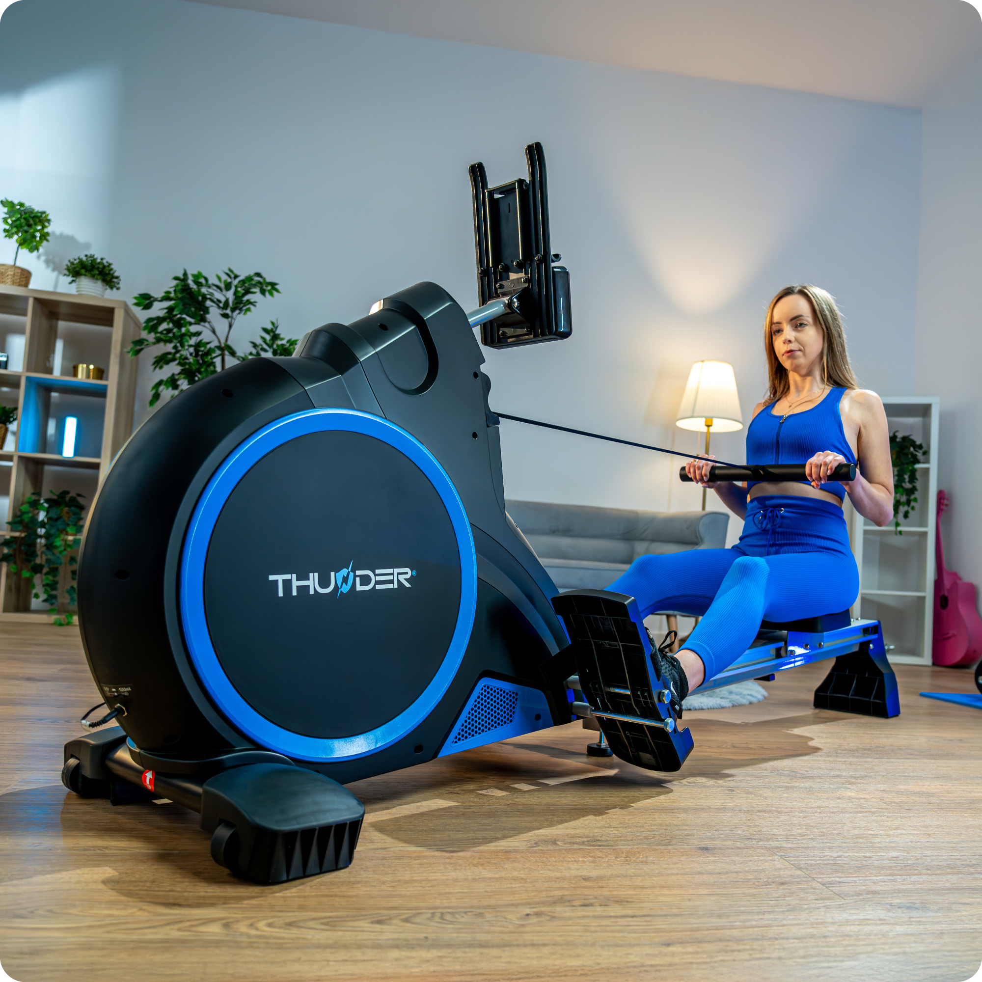 Kobieta w niebieskim stroju do ćwiczeń korzystająca z wioślarza. Ujęcie wnętrza z lampą i szafką.