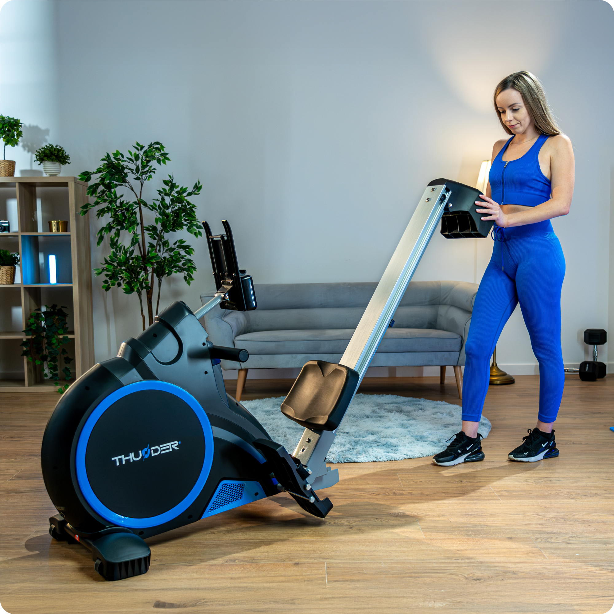 Kobieta korzystająca z wioślarza. Niebieskie legginsy i top. Tło: kanapa i roślina.