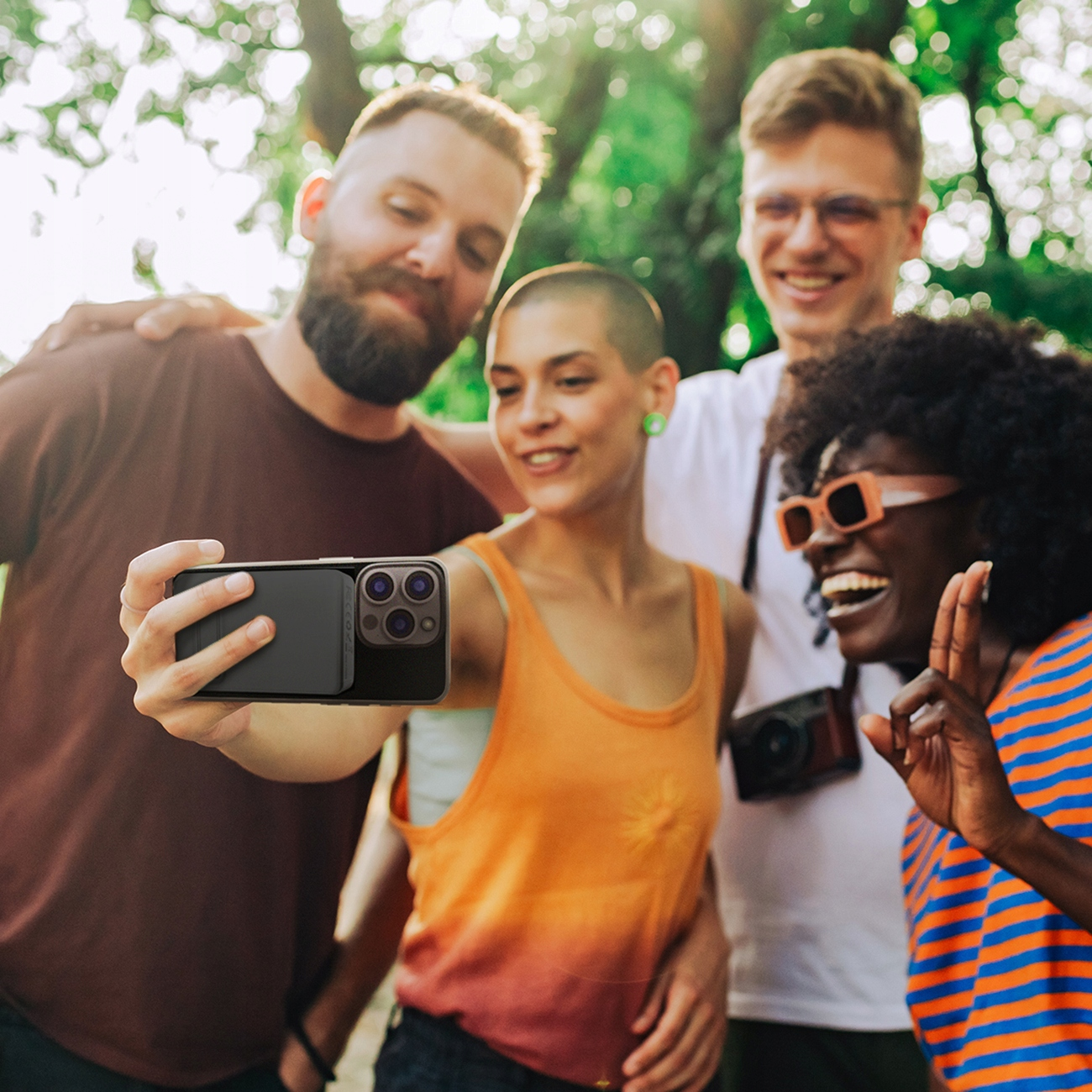 Czterech przyjaciół robiących selfie z etui na telefon. Słonecznie na zewnątrz z zielonymi liśćmi.