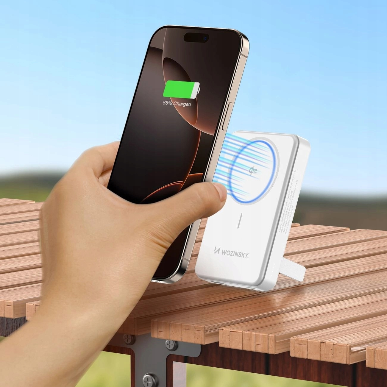 Ręka trzyma telefon ładujący się z białej ładowarki bezprzewodowej na drewnianym stole.
