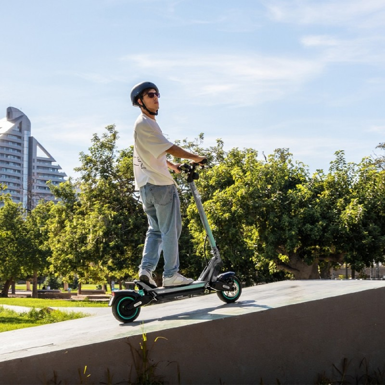 Un hombre montando un patinete eléctrico en una rampa, con árboles y edificios al fondo.