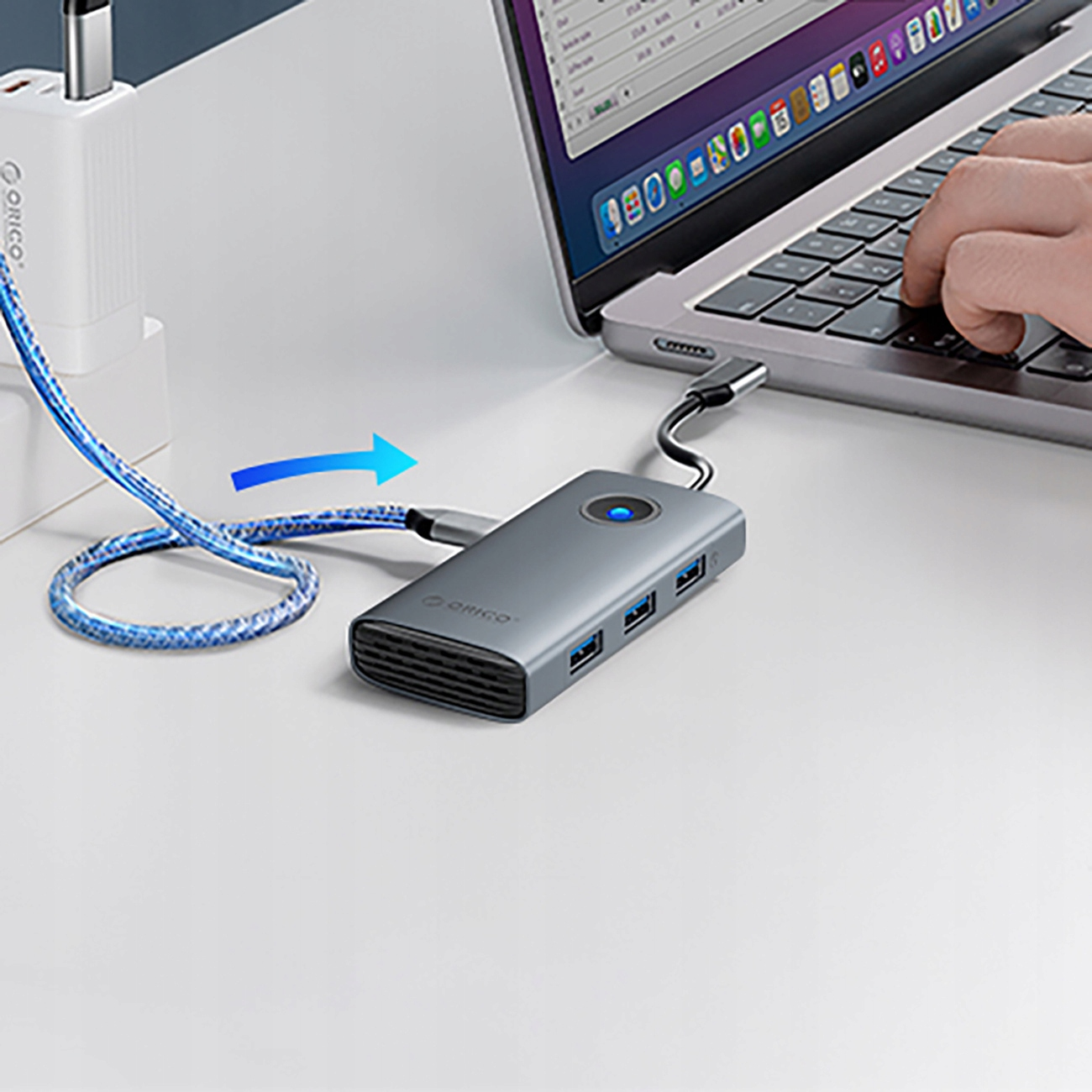 Srebrny hub USB podłączony do laptopa, z kablem ładującym i dłonią piszącą na klawiaturze.