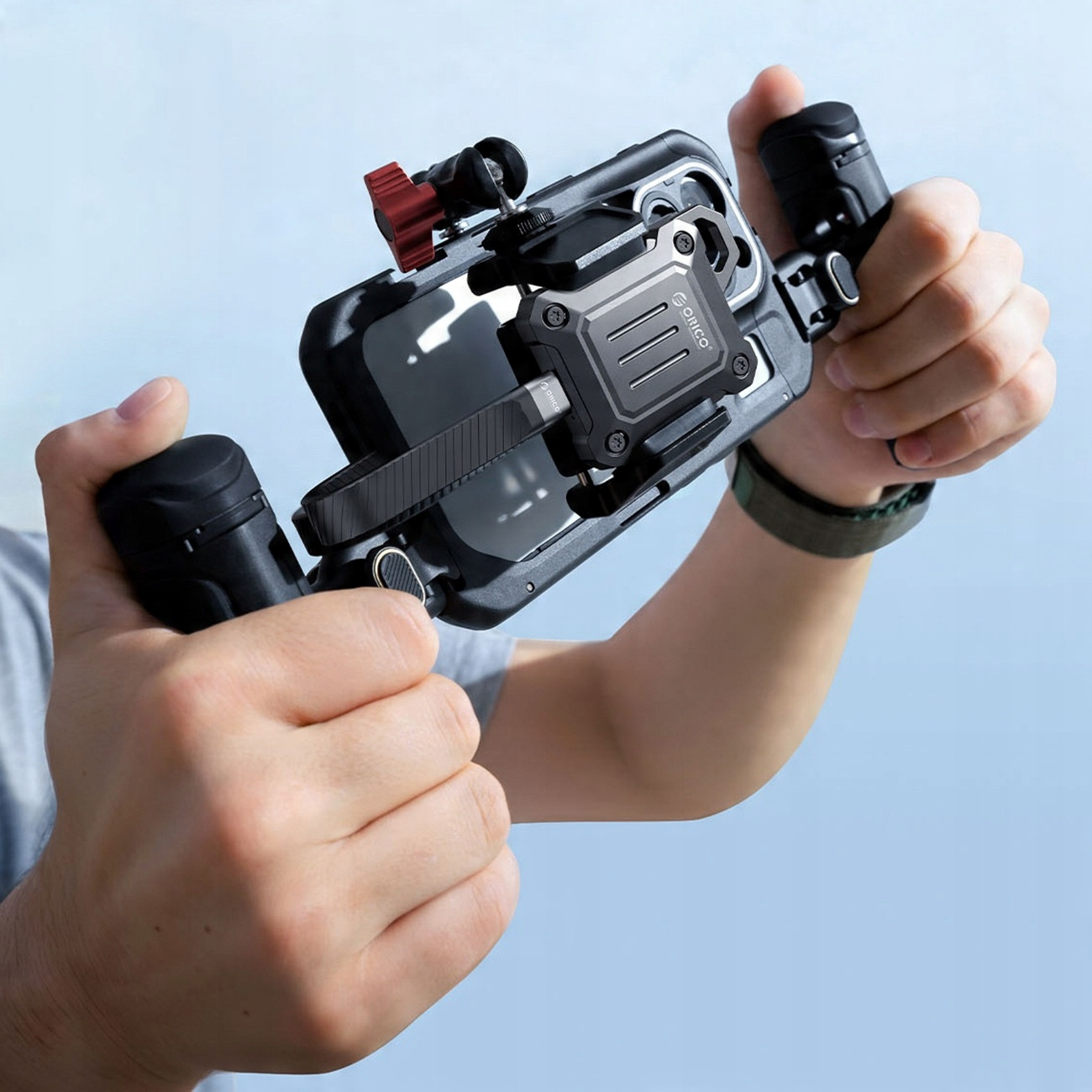 Dłonie trzymające czarne etui na telefon z dołączonymi akcesoriami na jasnoniebieskim tle.