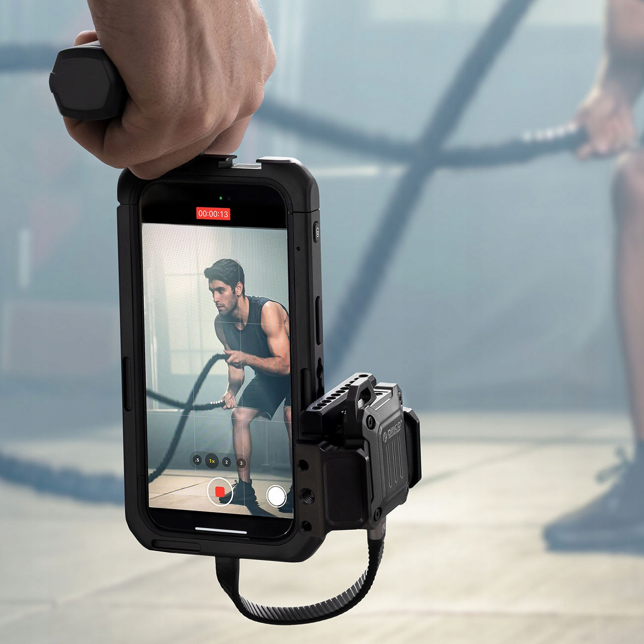 Ręka trzymająca telefon w etui, nagrywająca mężczyznę ćwiczącego z linami. Widoczna nazwa marki „ORICO”.