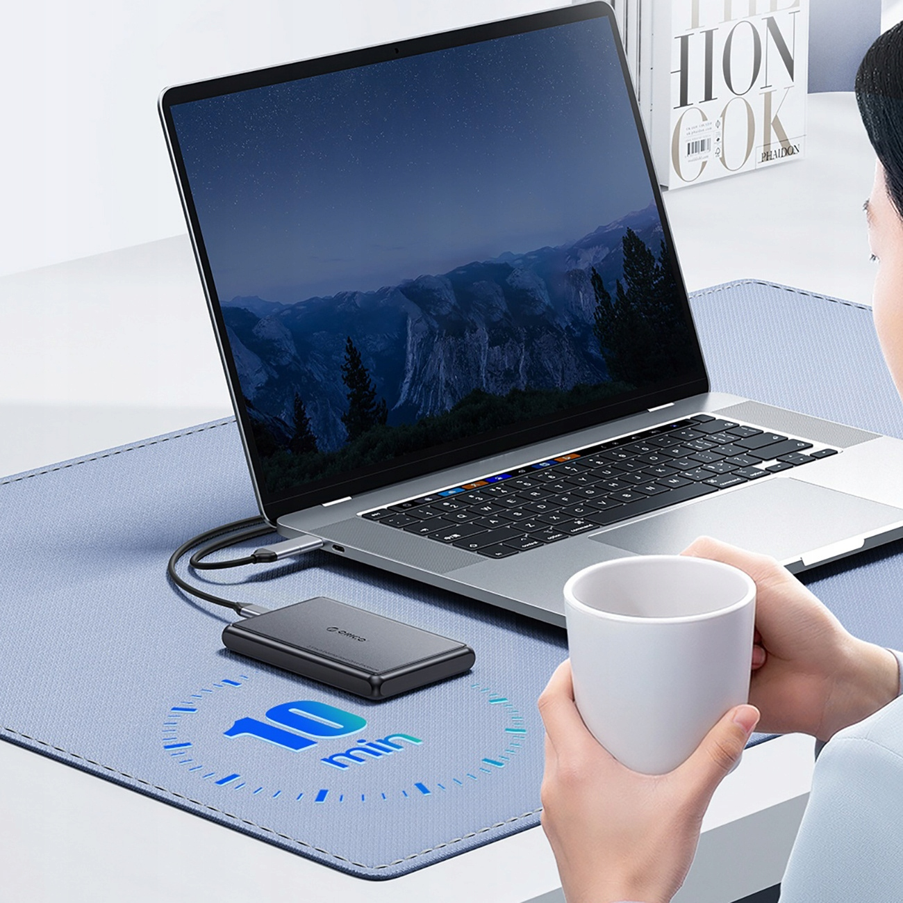 Laptop z podłączonym dyskiem zewnętrznym. Osoba trzyma biały kubek, tło: krajobraz górski.