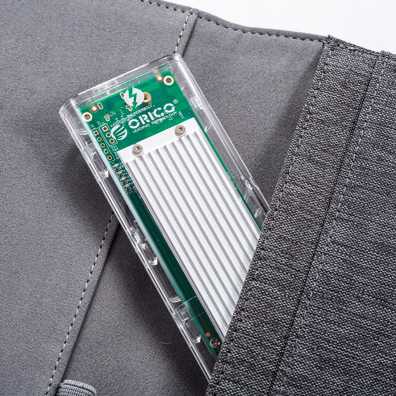Przezroczysty dysk SSD w szarej kieszeni. Marka Orico. Tło to szara tkanina.