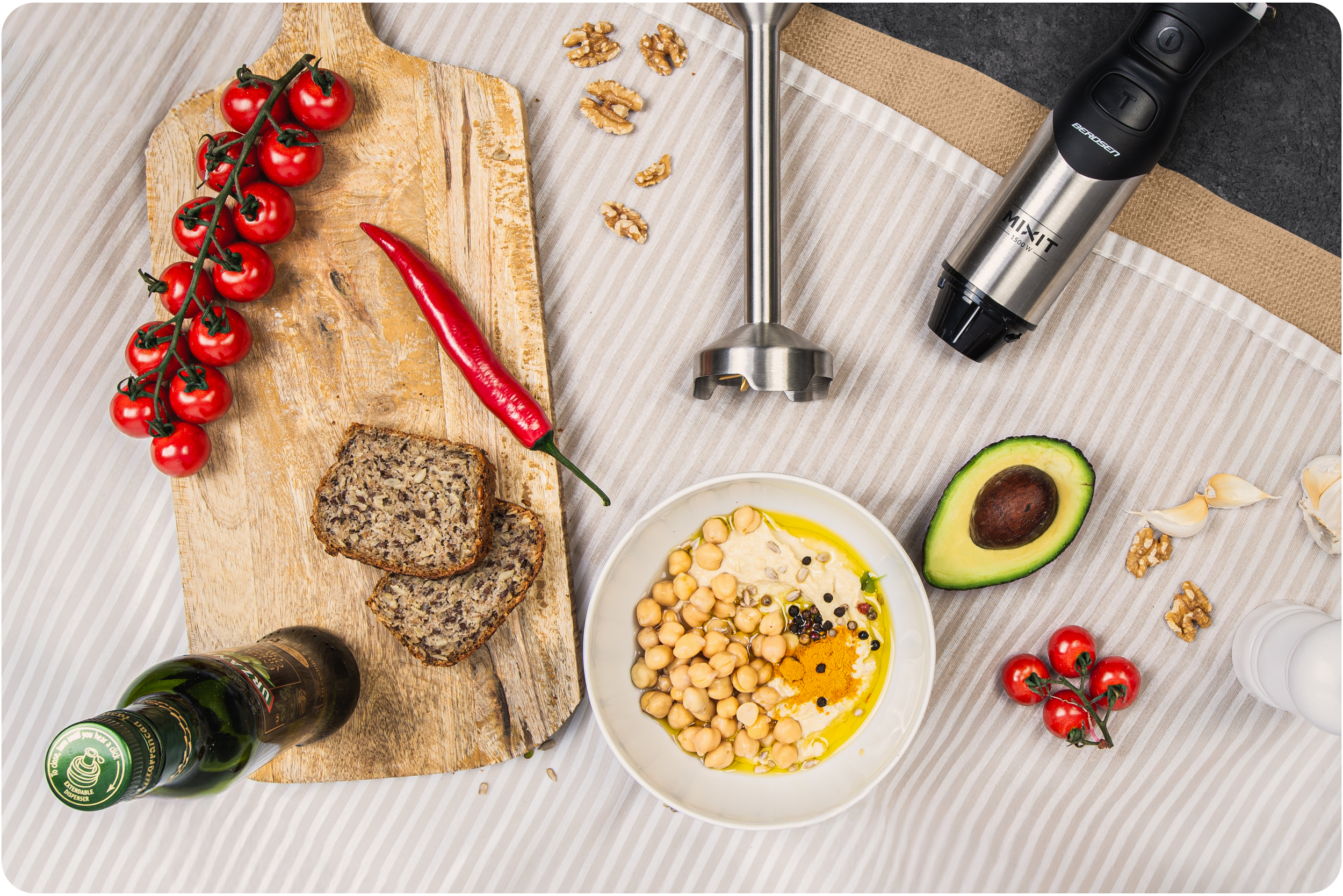 Ustawienie stołu z produktami spożywczymi, w tym blenderem ręcznym, humusem i pomidorami.