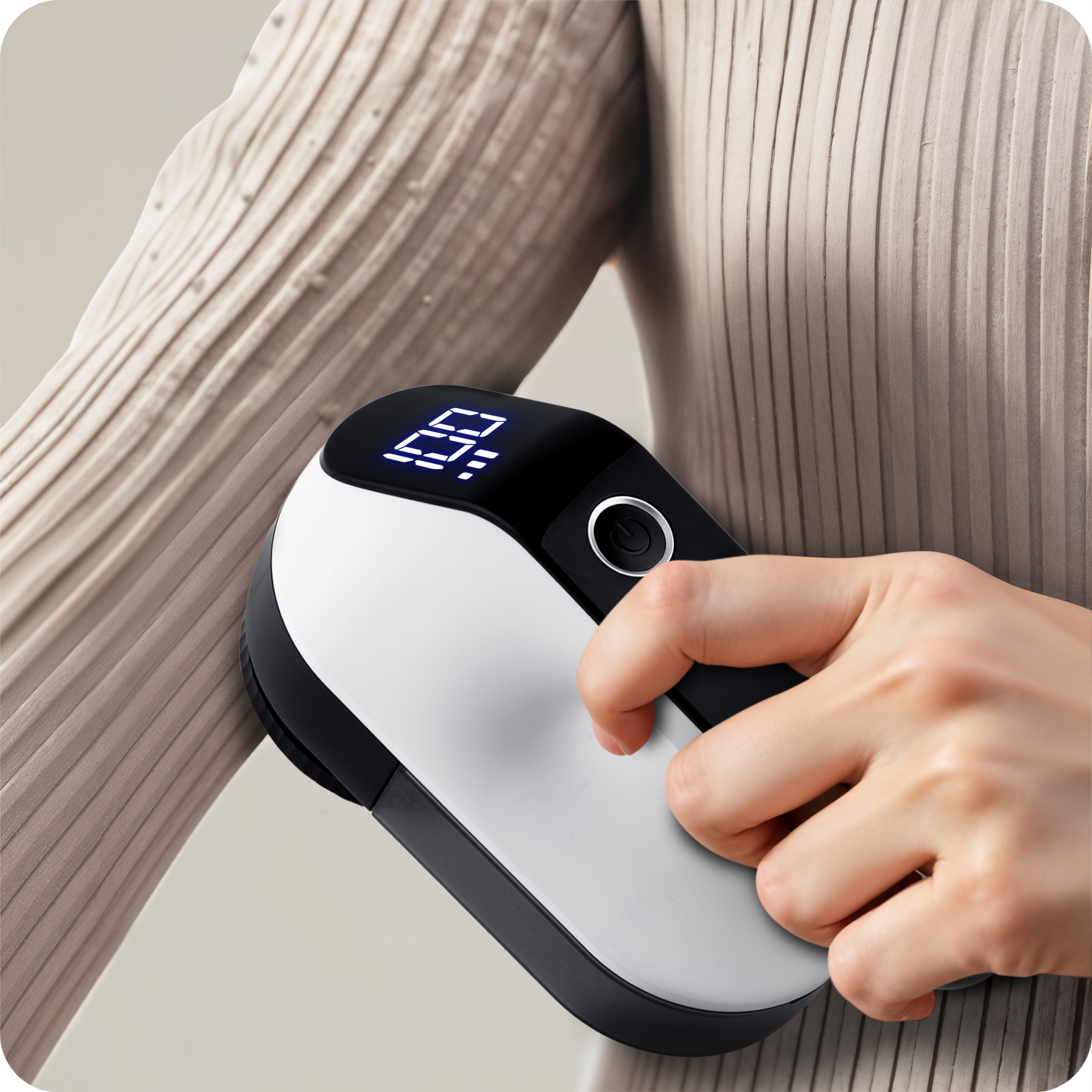 Osoba używa golarki do tkanin na beżowym swetrze. Golarka ma wyświetlacz i przycisk.