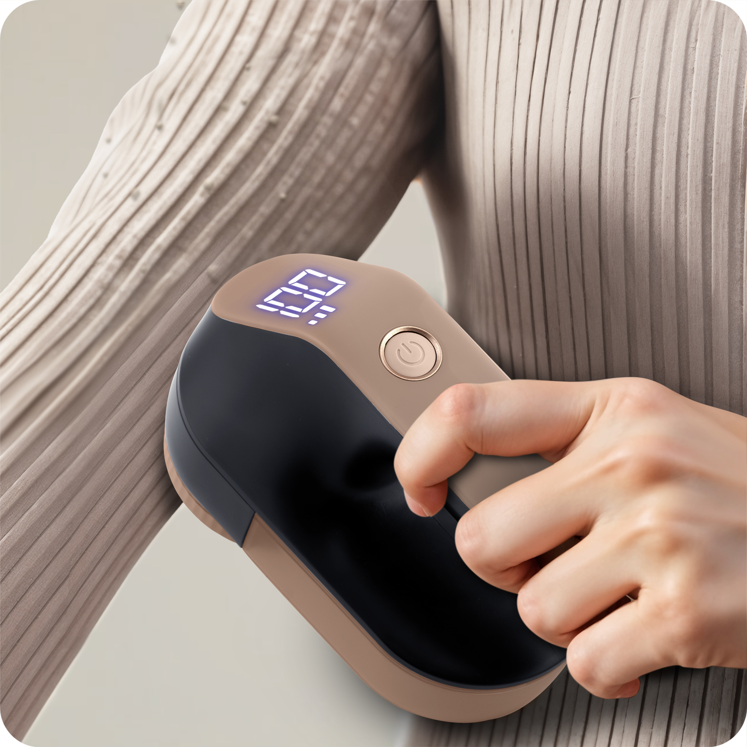 Dłoń trzyma urządzenie do usuwania kłaczków, z ekranem pokazującym 99. Urządzenie jest czarne i beżowe, na beżowej dzianinie.