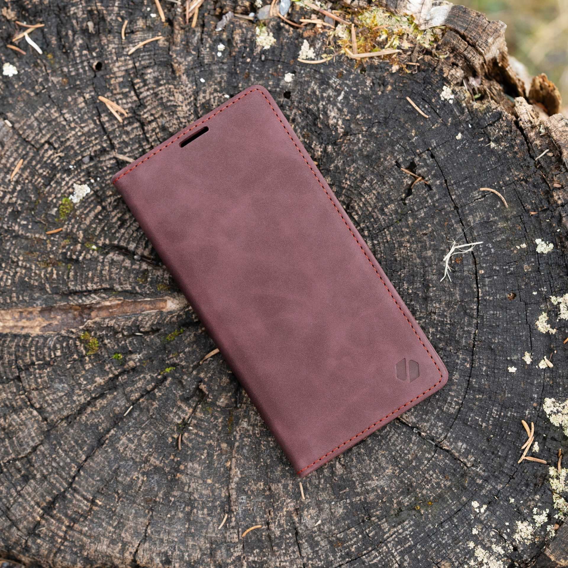 Brązowe etui na telefon leży na drewnianym pniu. Prostokątne, z logo i czerwonymi szwami.