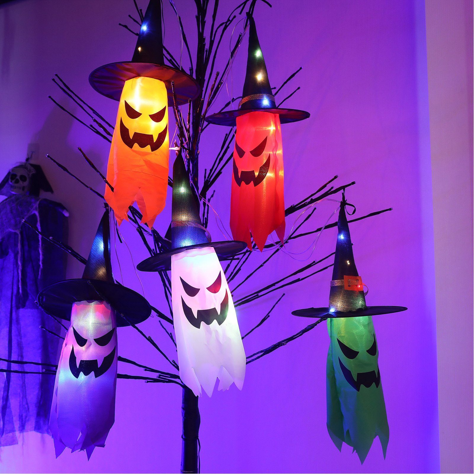 Halloween-Dekorationen mit Hexenhüten und Geistergesichtern hängen an einem Baum, beleuchtet.