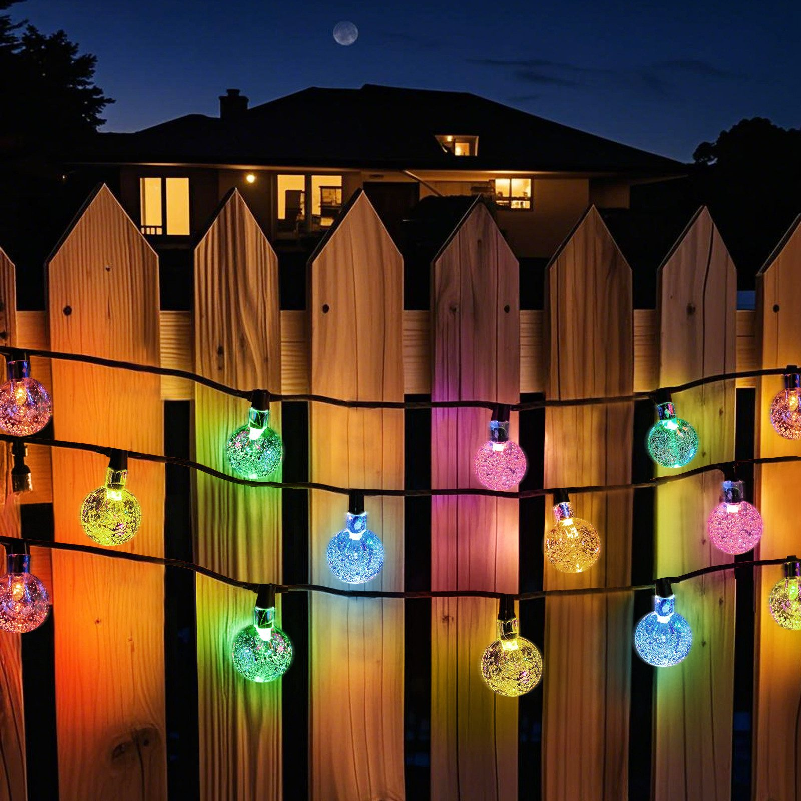 Lichterkette entlang eines Holzzauns bei Nacht. Die Lichter sind mehrfarbige Kugeln. Mond am Himmel sichtbar.
