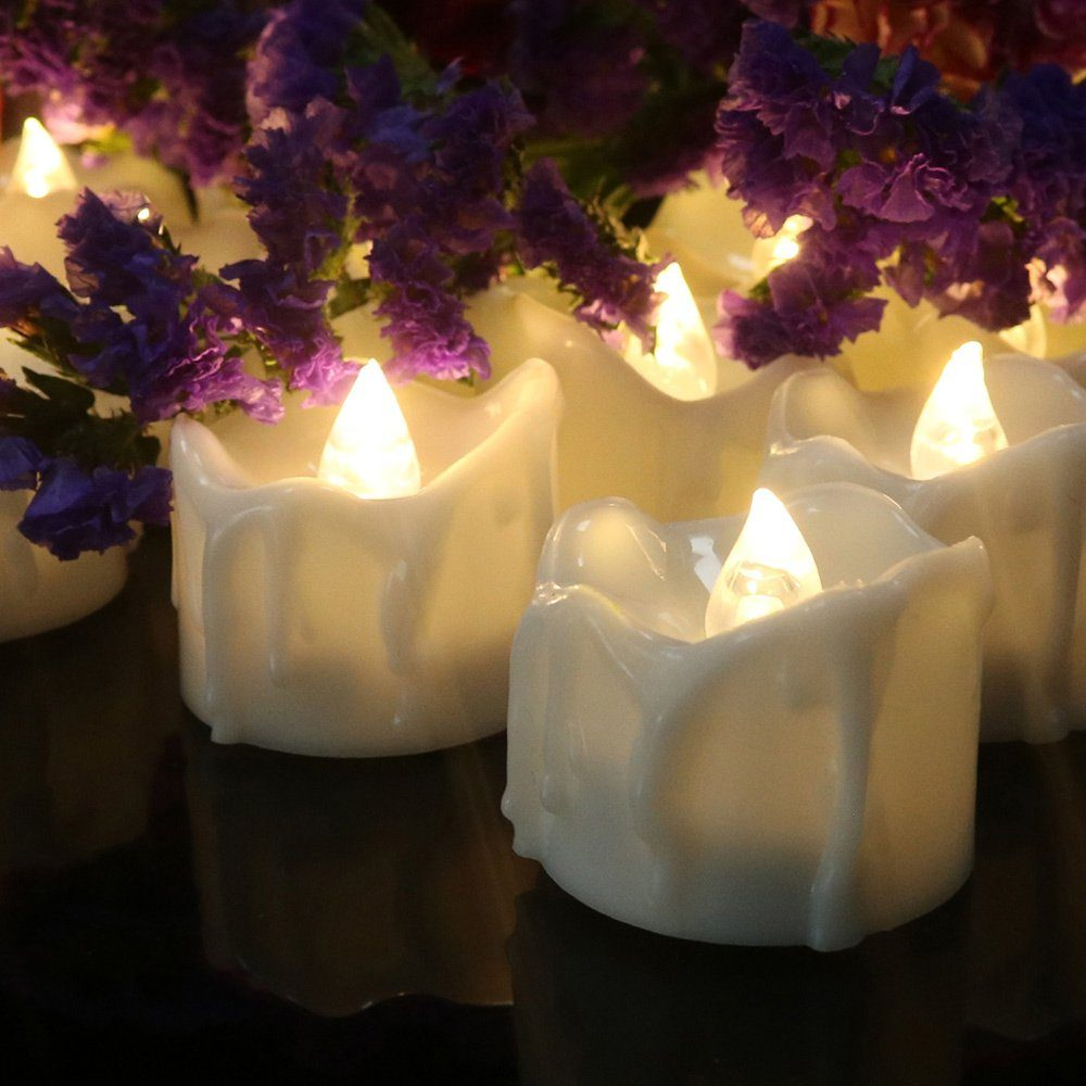 Mehrere brennende weiße Kerzen mit gelben Flammen. Violette Blumen sind im Hintergrund.