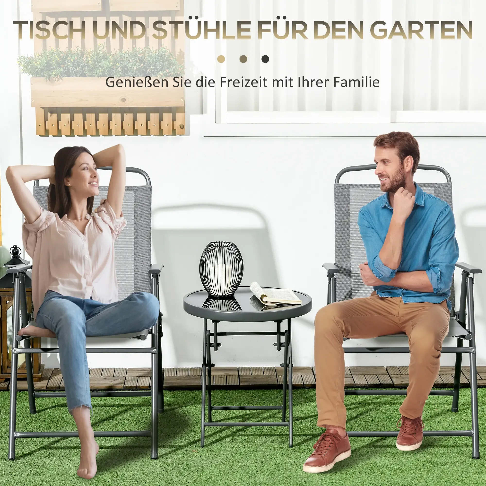 Ein Mann und eine Frau sitzen in grauen Stühlen an einem Tisch auf einer grünen Wiese.