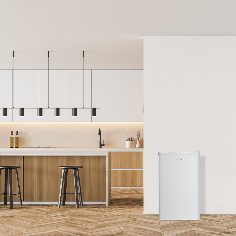 Un refrigerador blanco en una cocina moderna, junto a una pared blanca. Hay taburetes y luces en la escena.