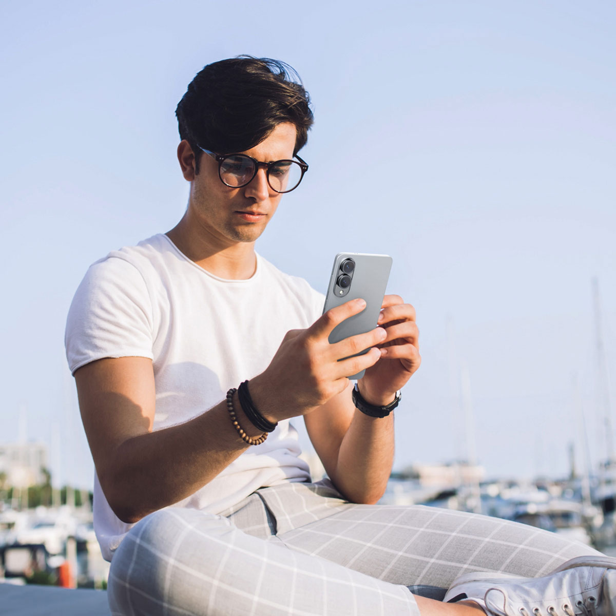 Un homme avec des lunettes, utilisant un téléphone, assis dehors avec des bateaux en arrière-plan.