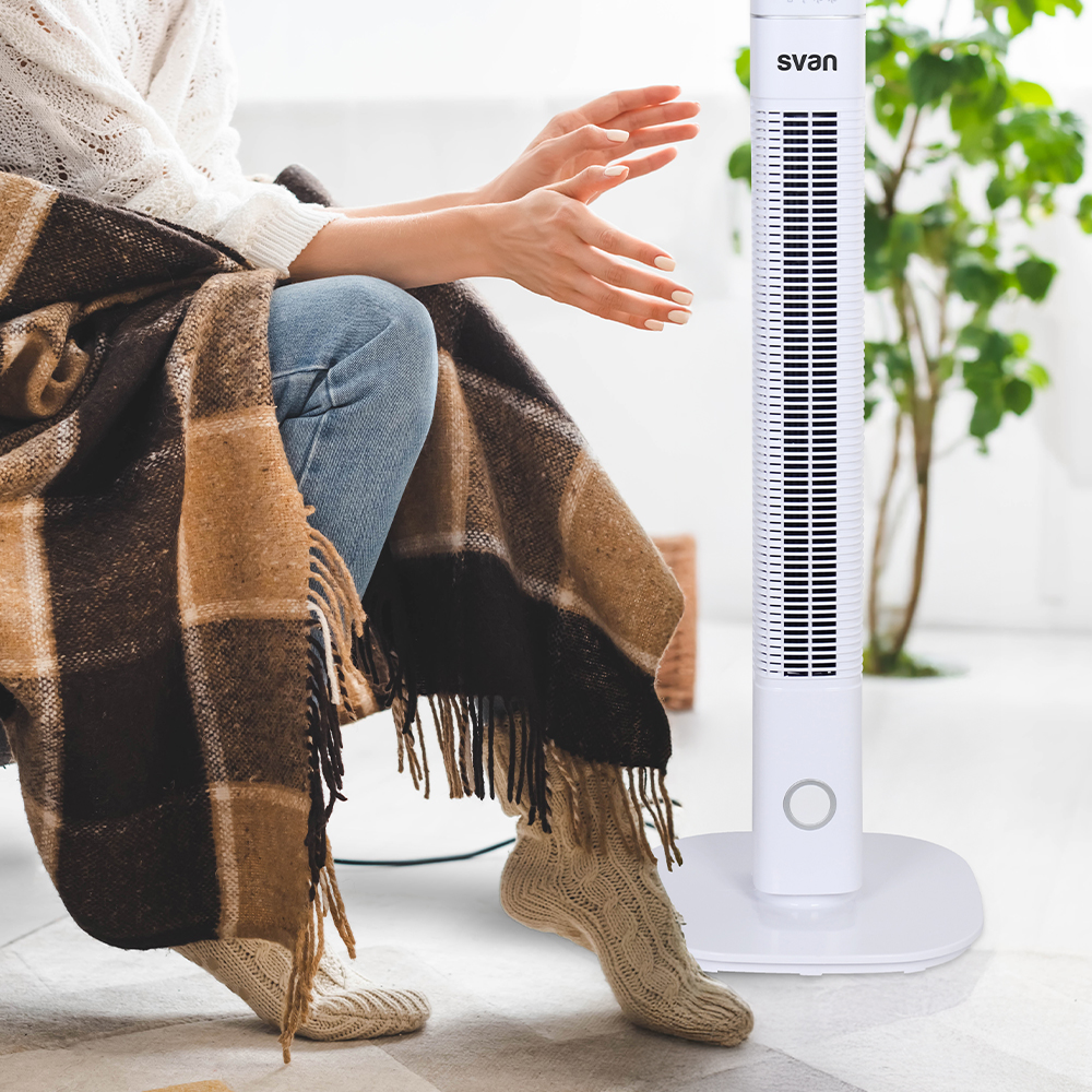 Una persona está sentada y extiende las manos hacia un ventilador blanco. Están cubiertos con una manta. El suelo es blanco.