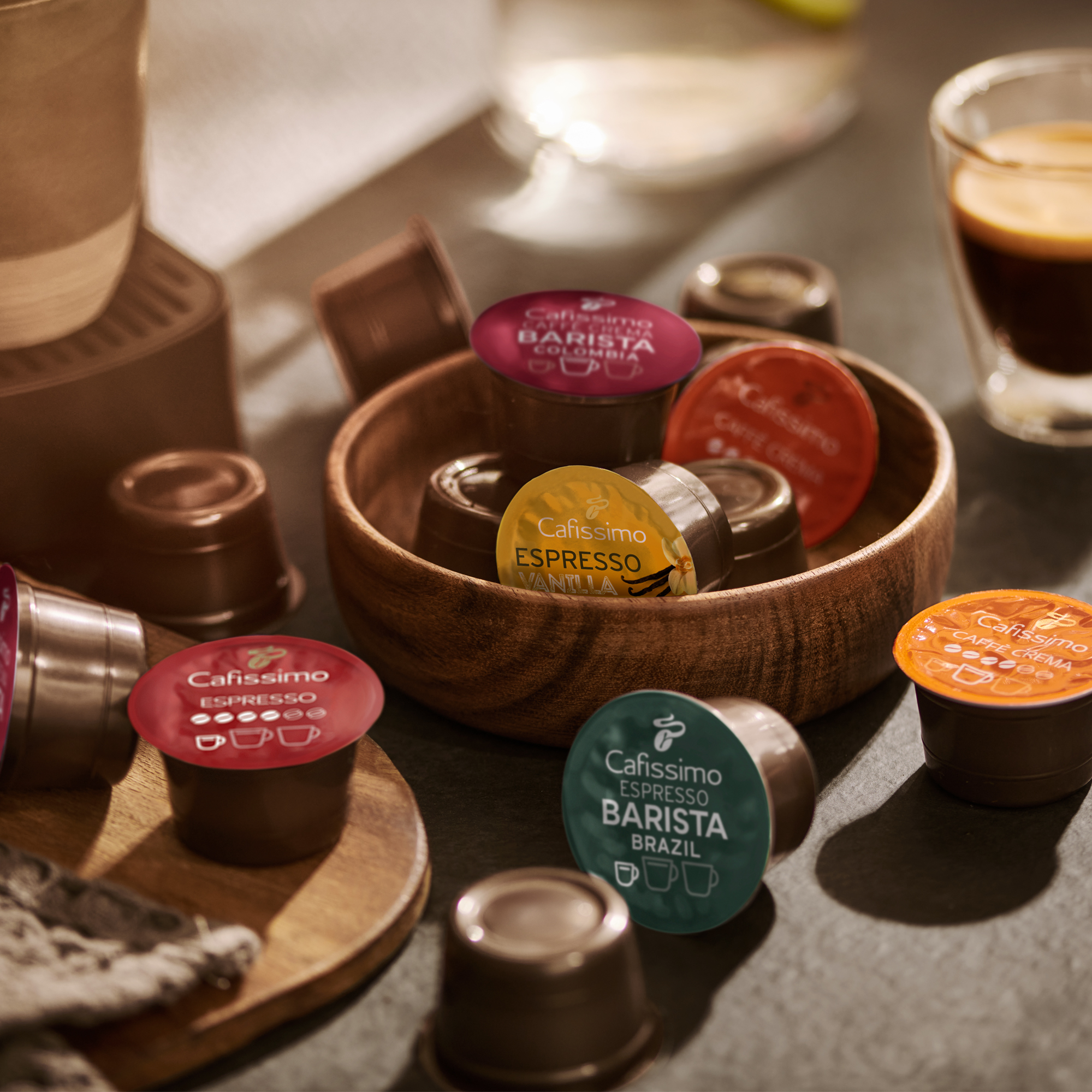 Kaffeepads in einer Holzschale, mit einer Tasse Kaffee und anderen Kaffeeartikeln. Verschiedene Geschmacksrichtungen sind sichtbar.