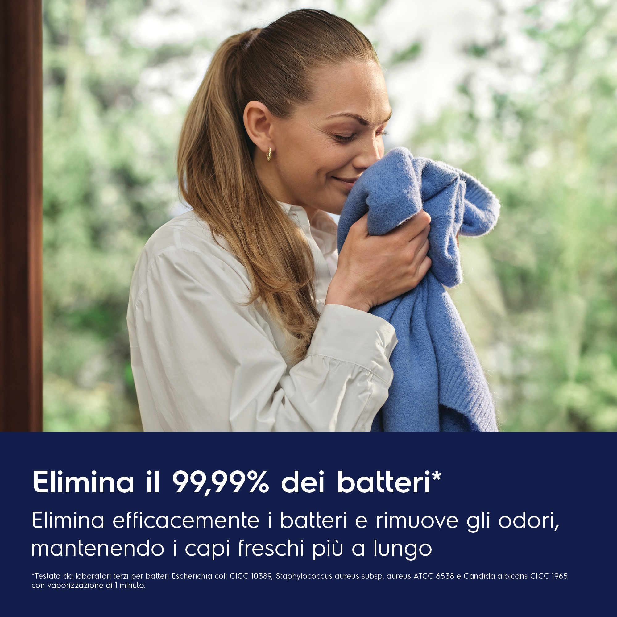 Donna che annusa un maglione blu, indossa una camicia bianca e sorride in casa.