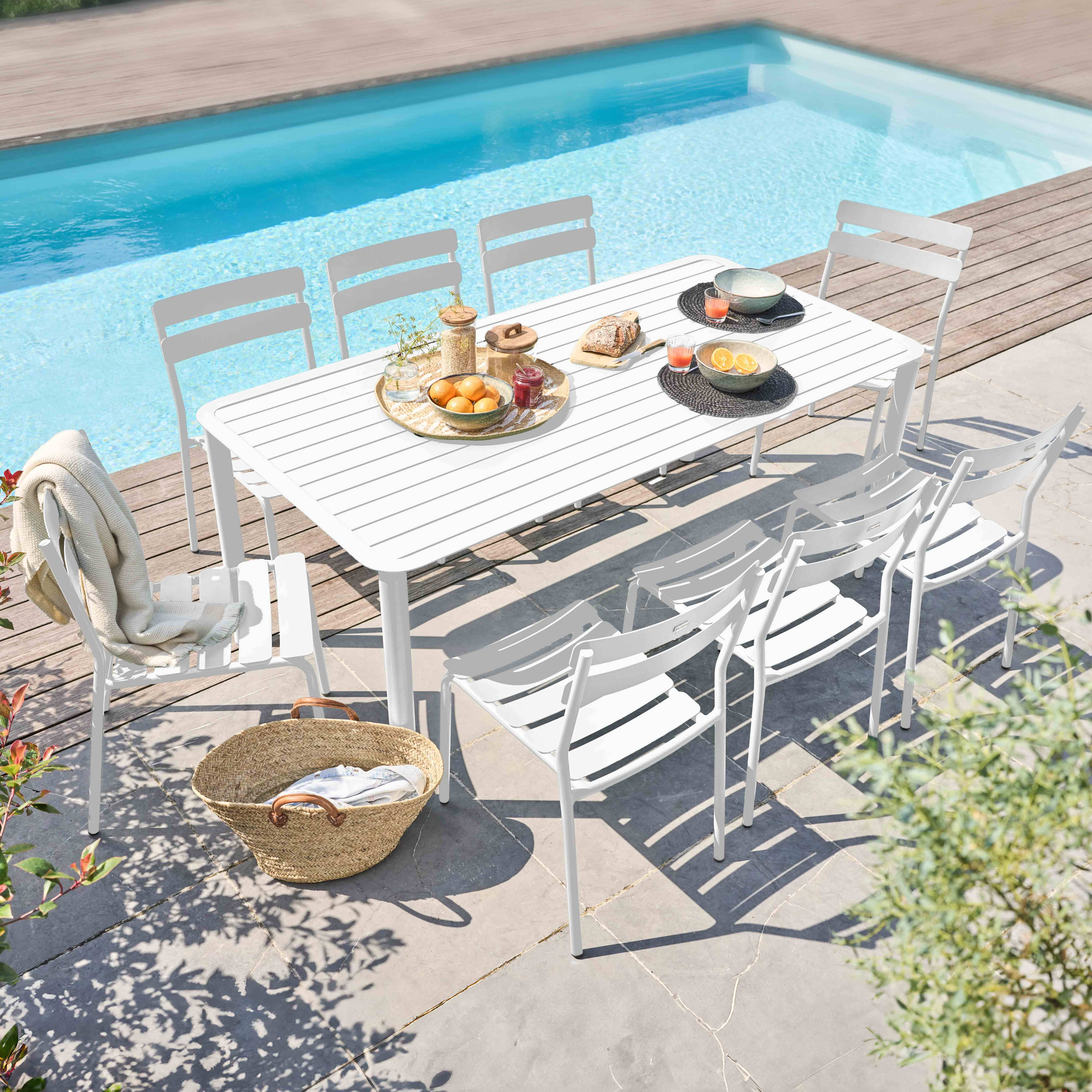 Ein langer weißer Tisch und acht weiße Stühle stehen neben einem blauen Swimmingpool.
