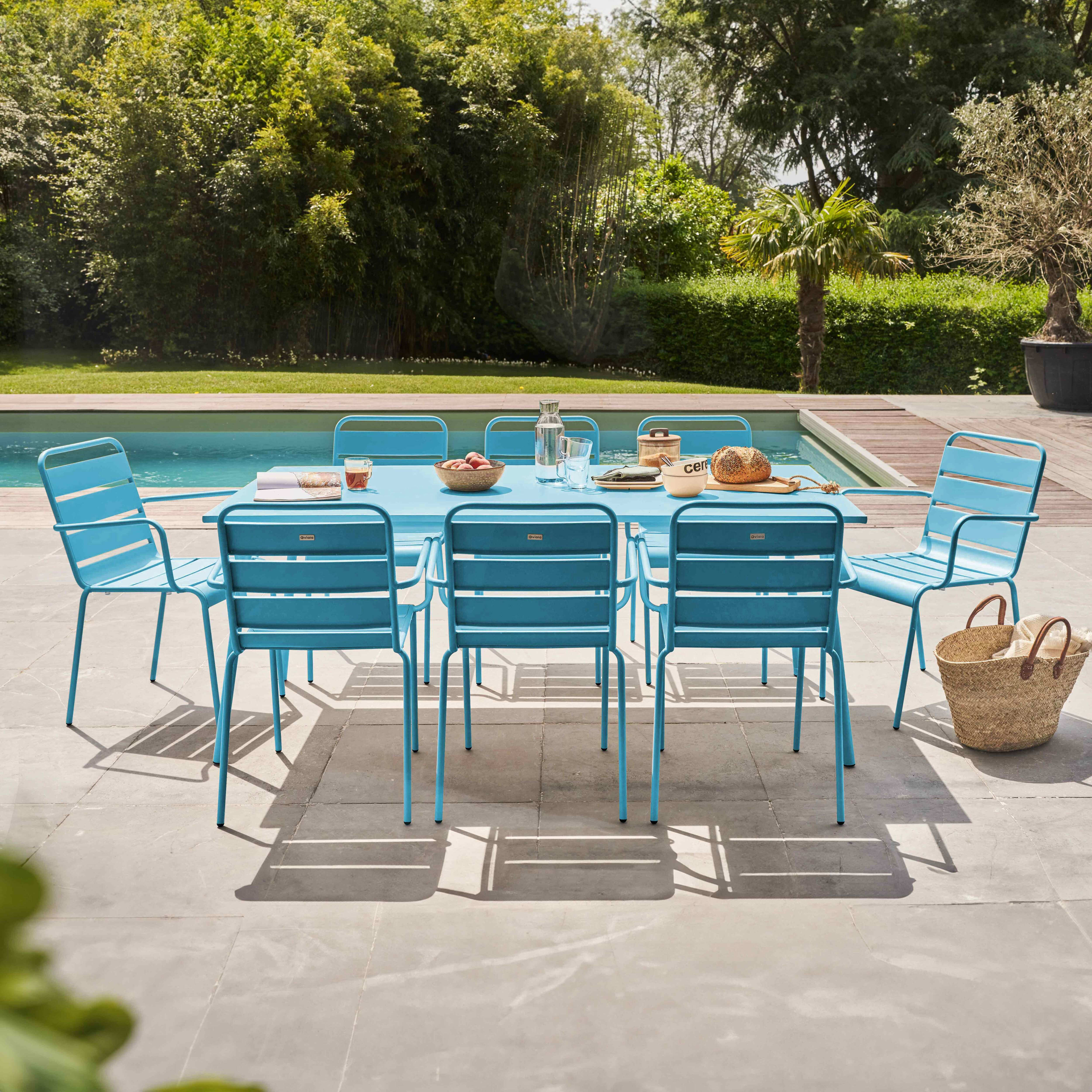 Ess-Set im Freien mit blauem Tisch und Stühlen. Auf einer Terrasse mit Pool und Bäumen im Hintergrund.