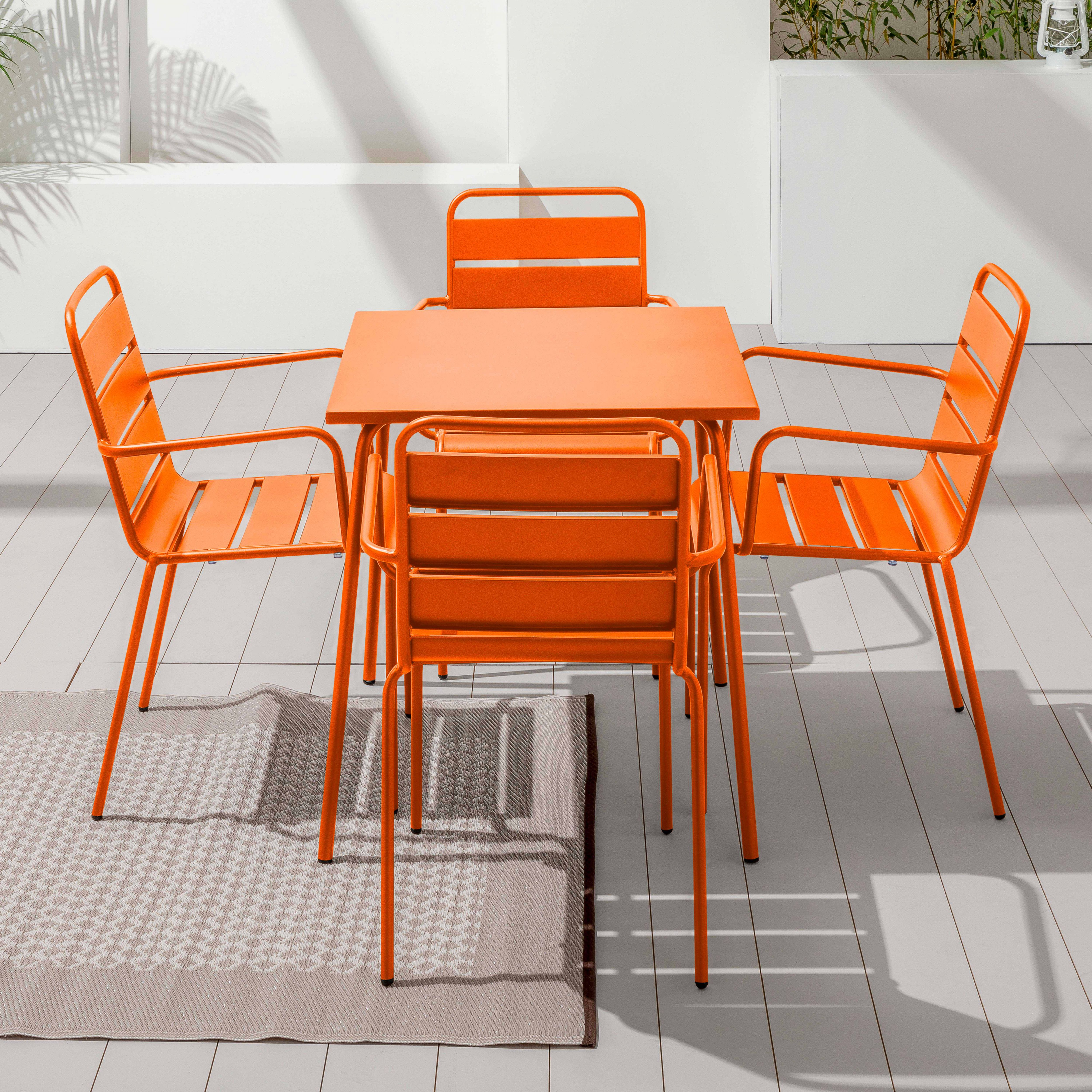 Ein orangefarbenes Ess-Set im Freien mit einem quadratischen Tisch, vier Stühlen und einem Teppich. Die Szene befindet sich auf einem Holzdeck.