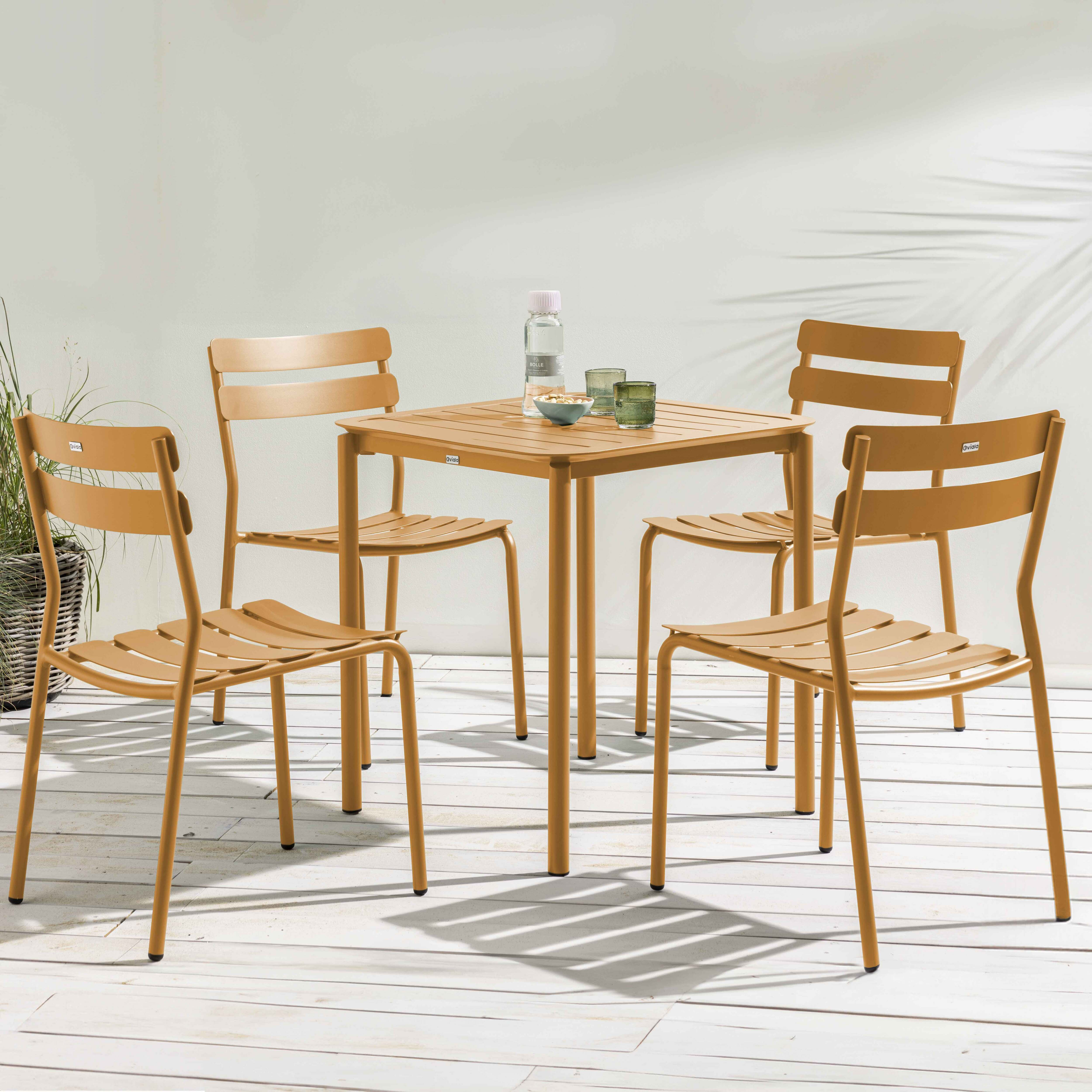 Essgruppe im Freien mit einem quadratischen Tisch und vier Stühlen. Das Set ist hellbraun und befindet sich auf einer weißen Holzterrasse.