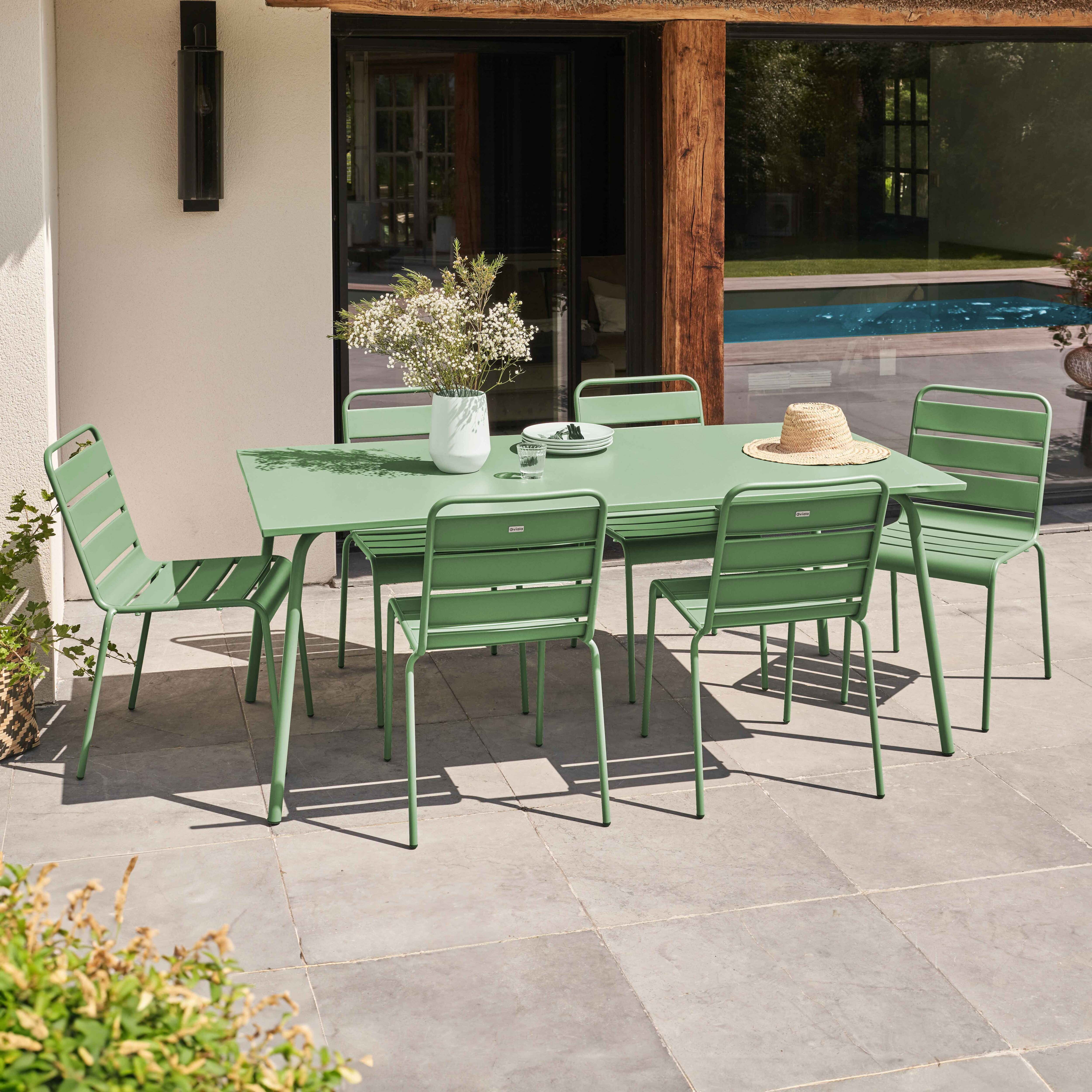 Grüner Tisch und Stühle auf einer Terrasse. Weiße Vase mit Blumen. Pool und Bäume im Hintergrund.