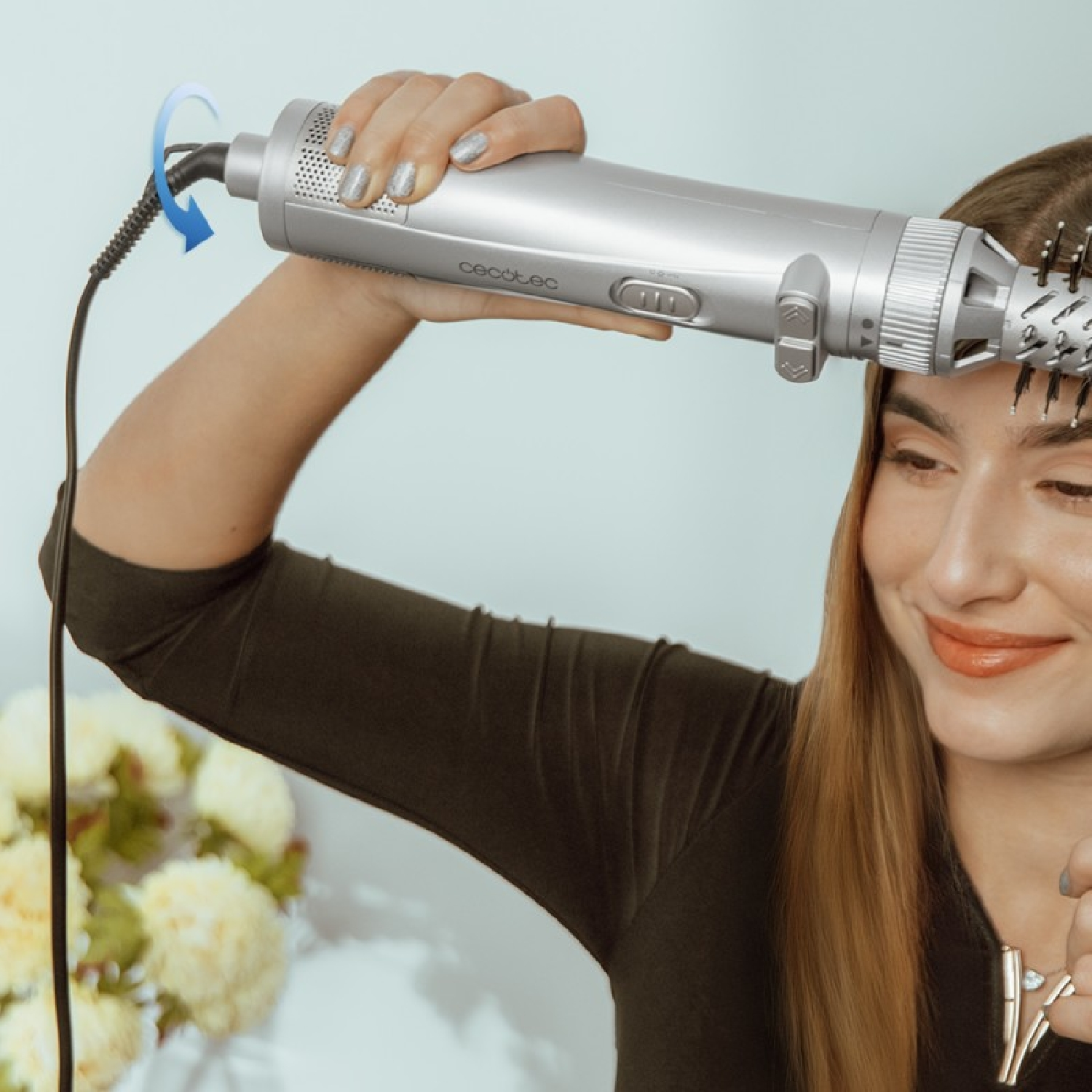 Una donna tiene in mano uno strumento per lo styling dei capelli, sorridendo, con fiori sullo sfondo.
