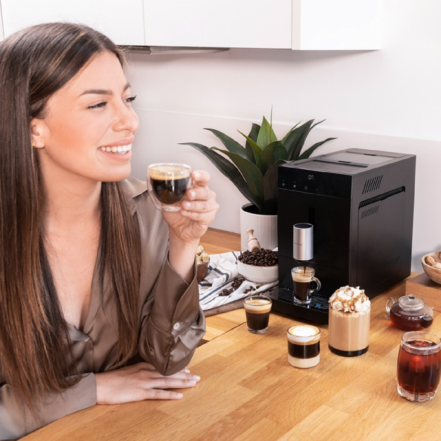 Donna beve caffè vicino a una macchina da caffè nera, con tazze e una pianta sul tavolo.