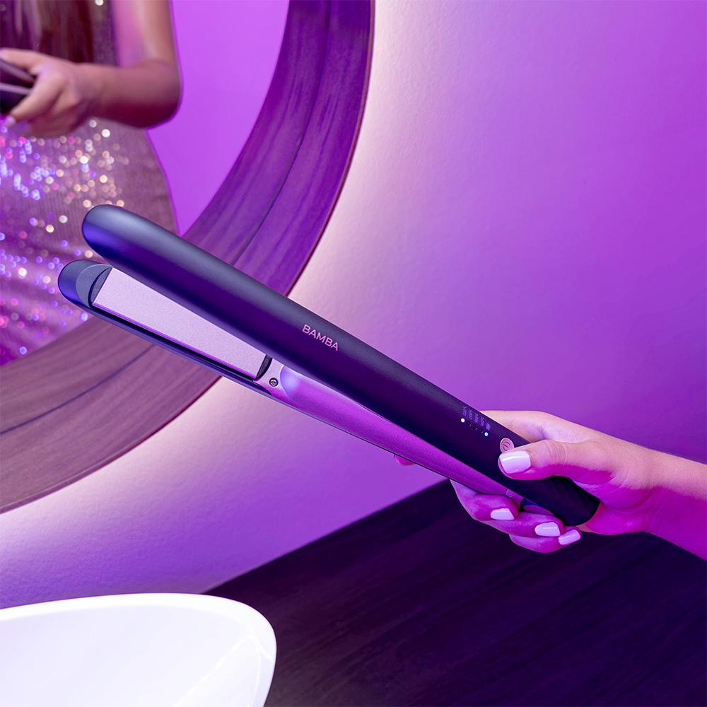 Ręka trzyma czarno-srebrną prostownicę do włosów przed lustrem z fioletowym oświetleniem.