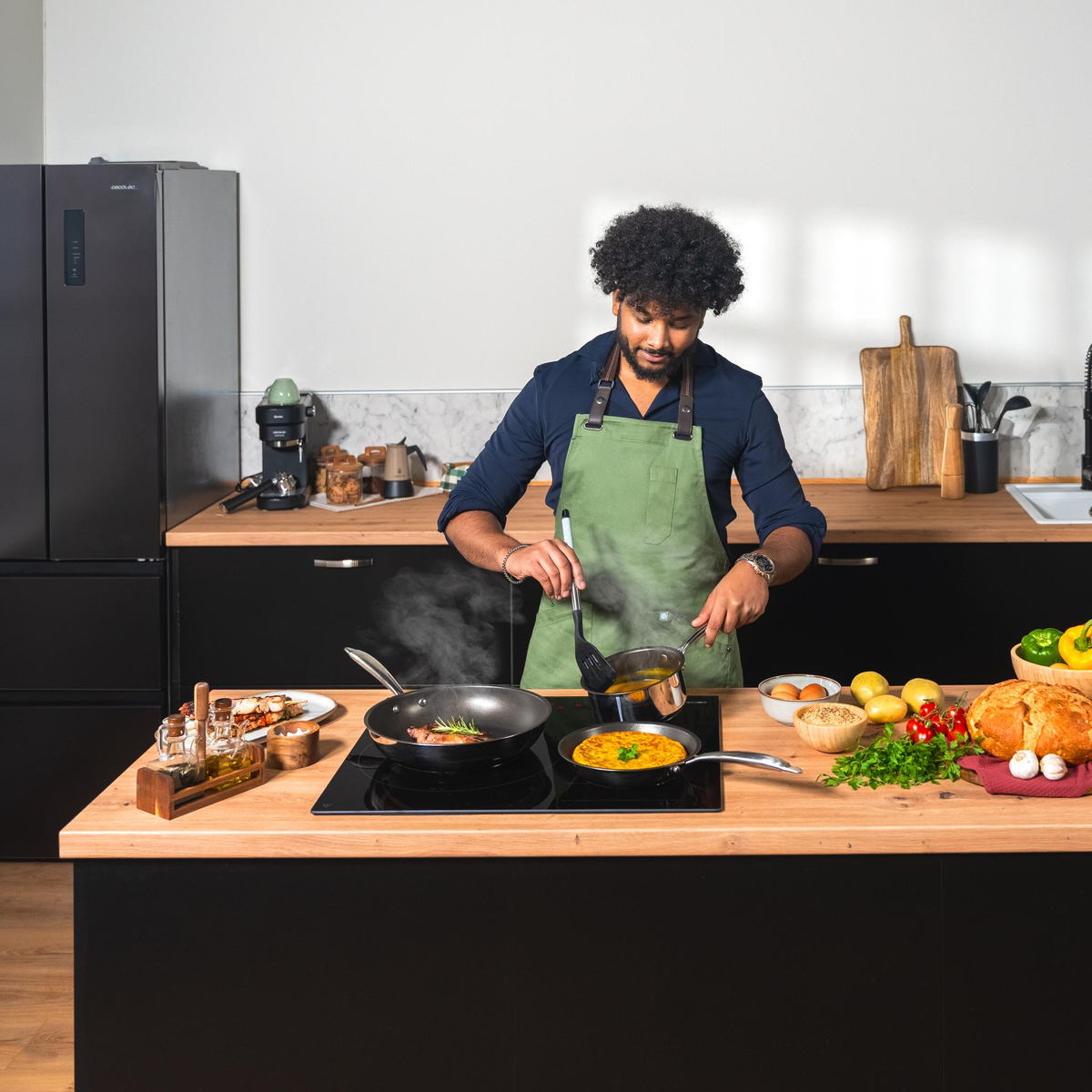 Mężczyzna gotuje na czarnej płycie z różnymi składnikami i daniami. W tle lodówka.
