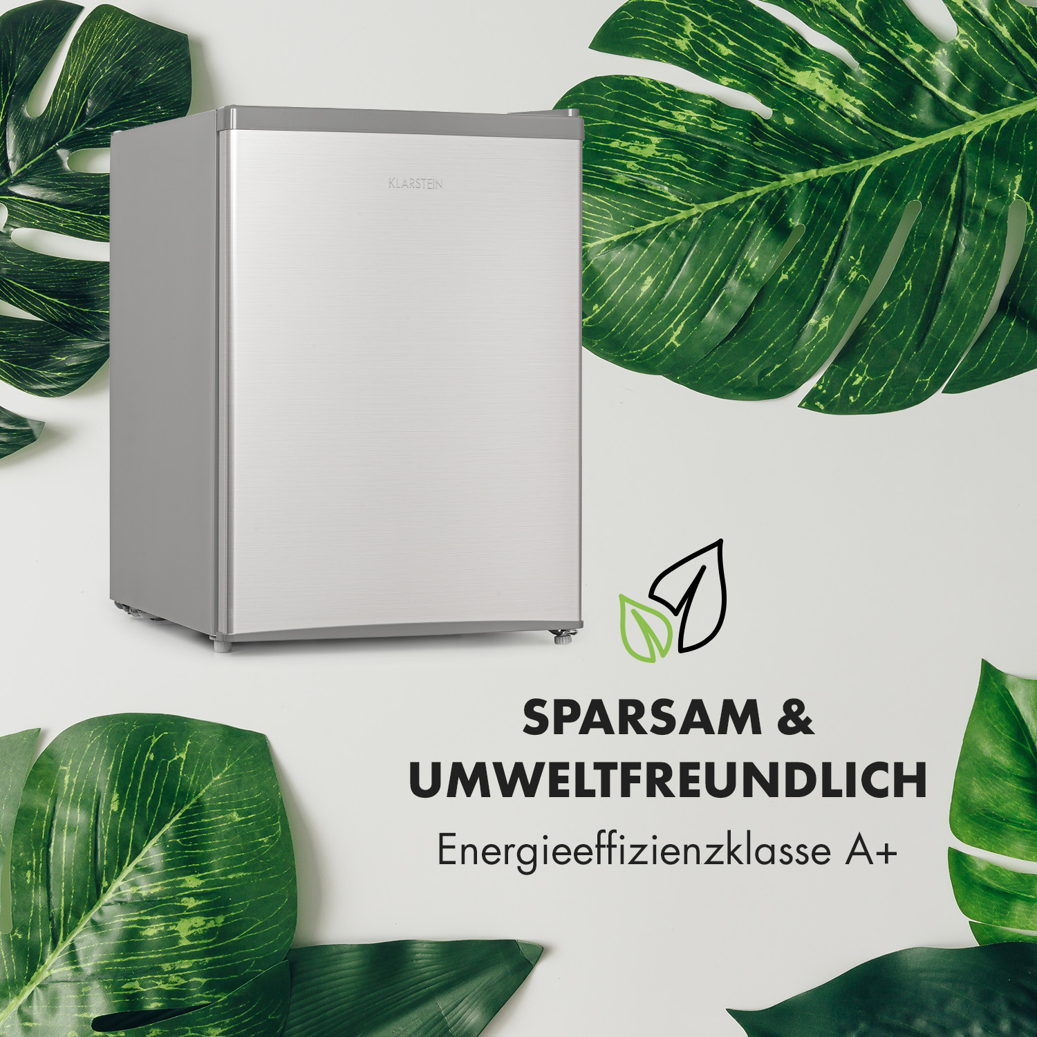 Ein silberner Mini-Kühlschrank steht vor einer weißen Wand, umgeben von grünen Blättern.