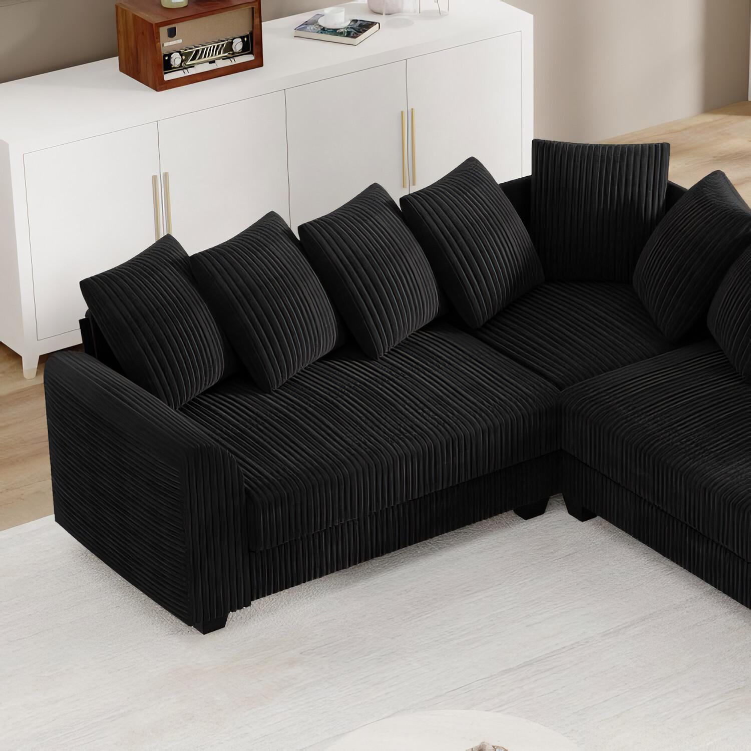 Ein schwarzes Ecksofa mit mehreren Kissen auf einem weißen Teppich. Weißer Schrank im Hintergrund.