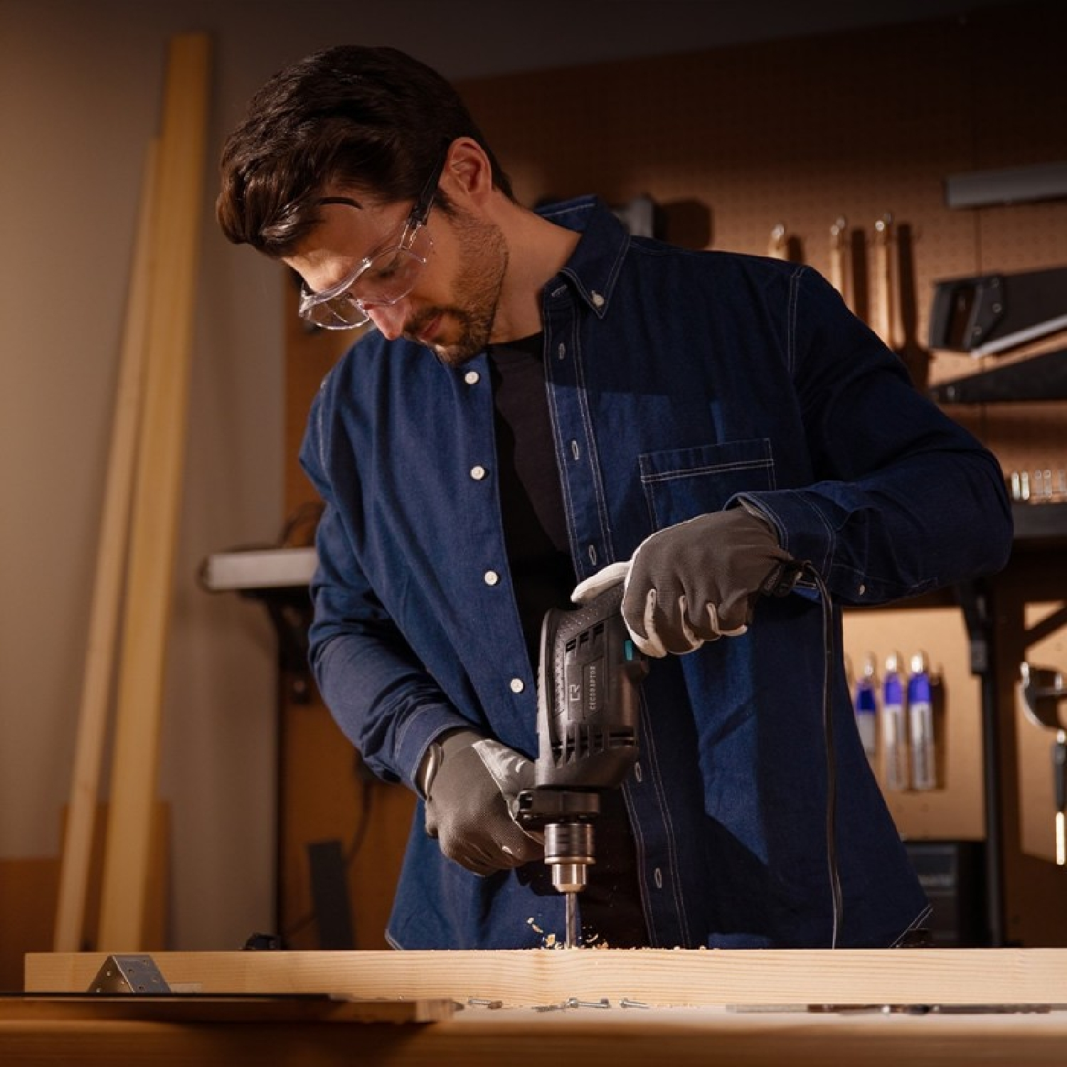 Mann mit Schutzbrille bohrt in Holz. Bohrer und Holz sind fokussiert.