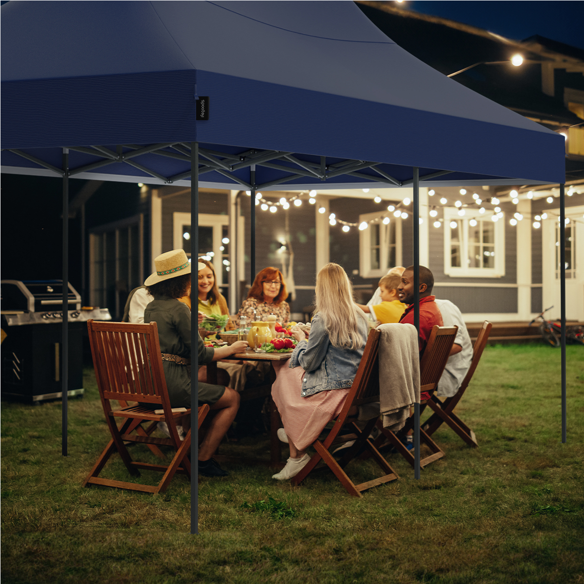 A group of people are seated at a table under a party tent at night with string lights.