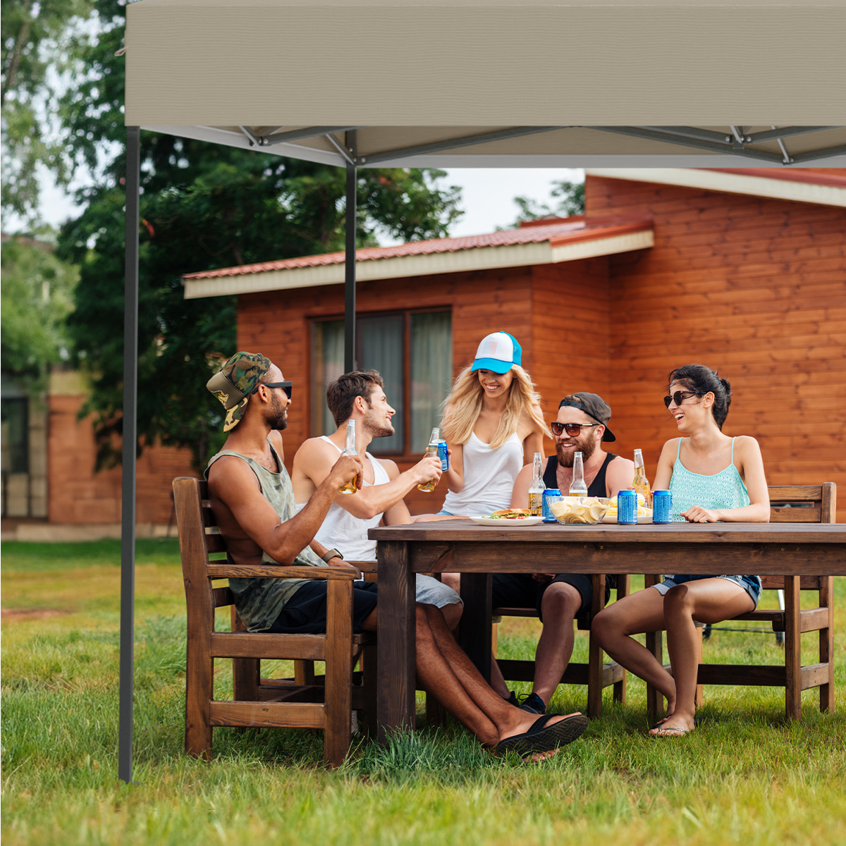 Five people under a canopy, enjoying drinks and socializing. Wooden table and chairs on grass, a house in the background.