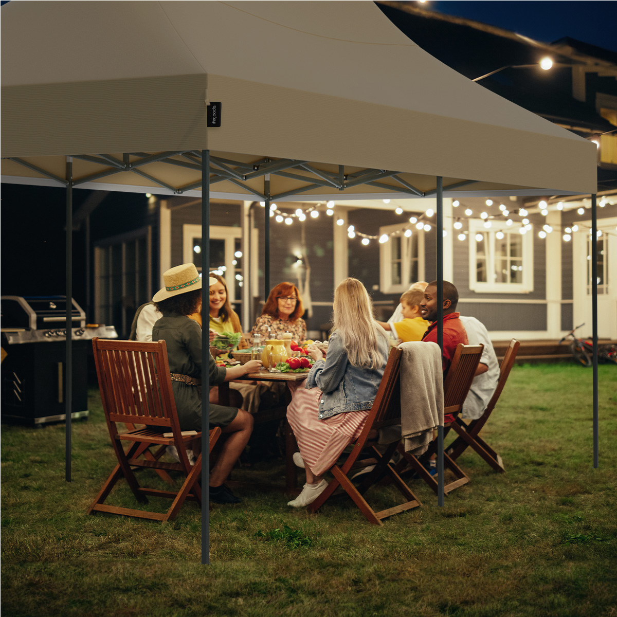 A group of people are seated at a table under a party tent at night with string lights.