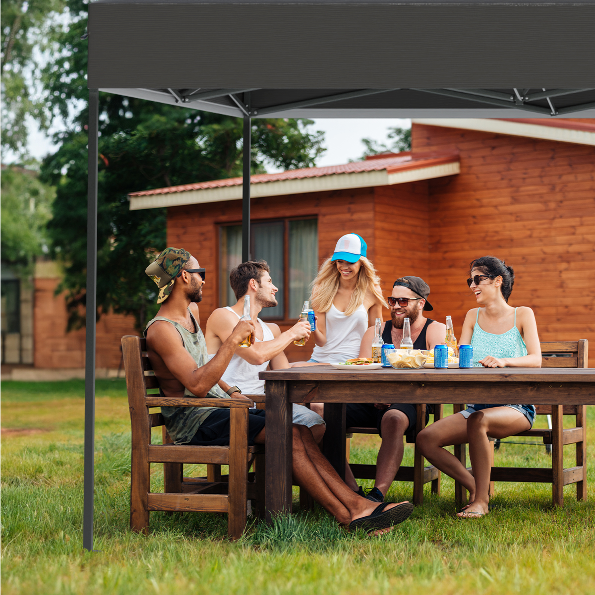 Five people sit at a table under a grey canopy, enjoying drinks. A wooden house is in the background.