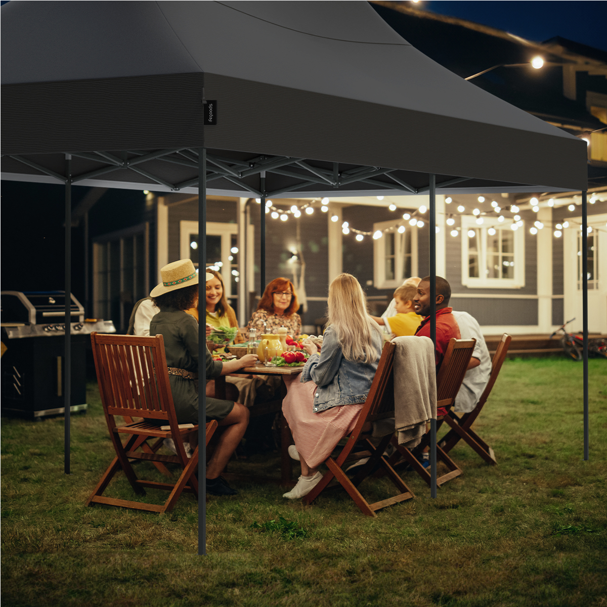 Group of people at a table under a black tent at night, with string lights and a house in the background.