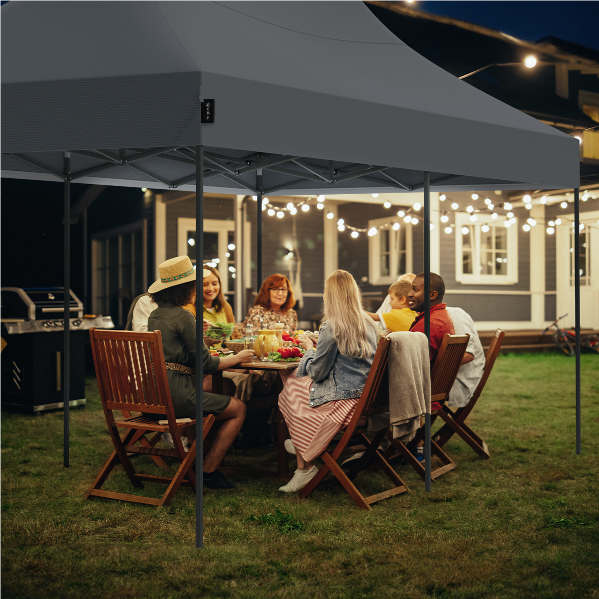 A group of people are eating under a grey canopy tent at night, with lights strung around.