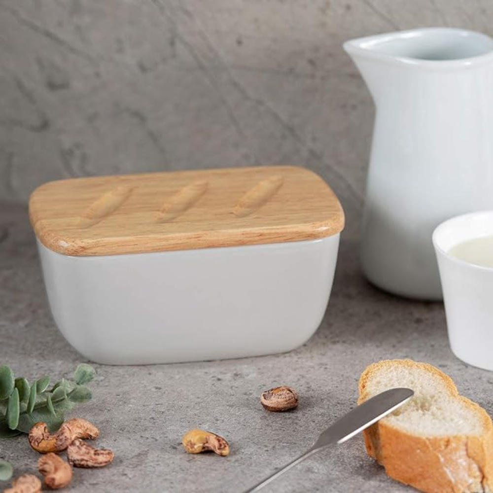 Una burriera bianca con coperchio in legno e pane e noci sullo sfondo.