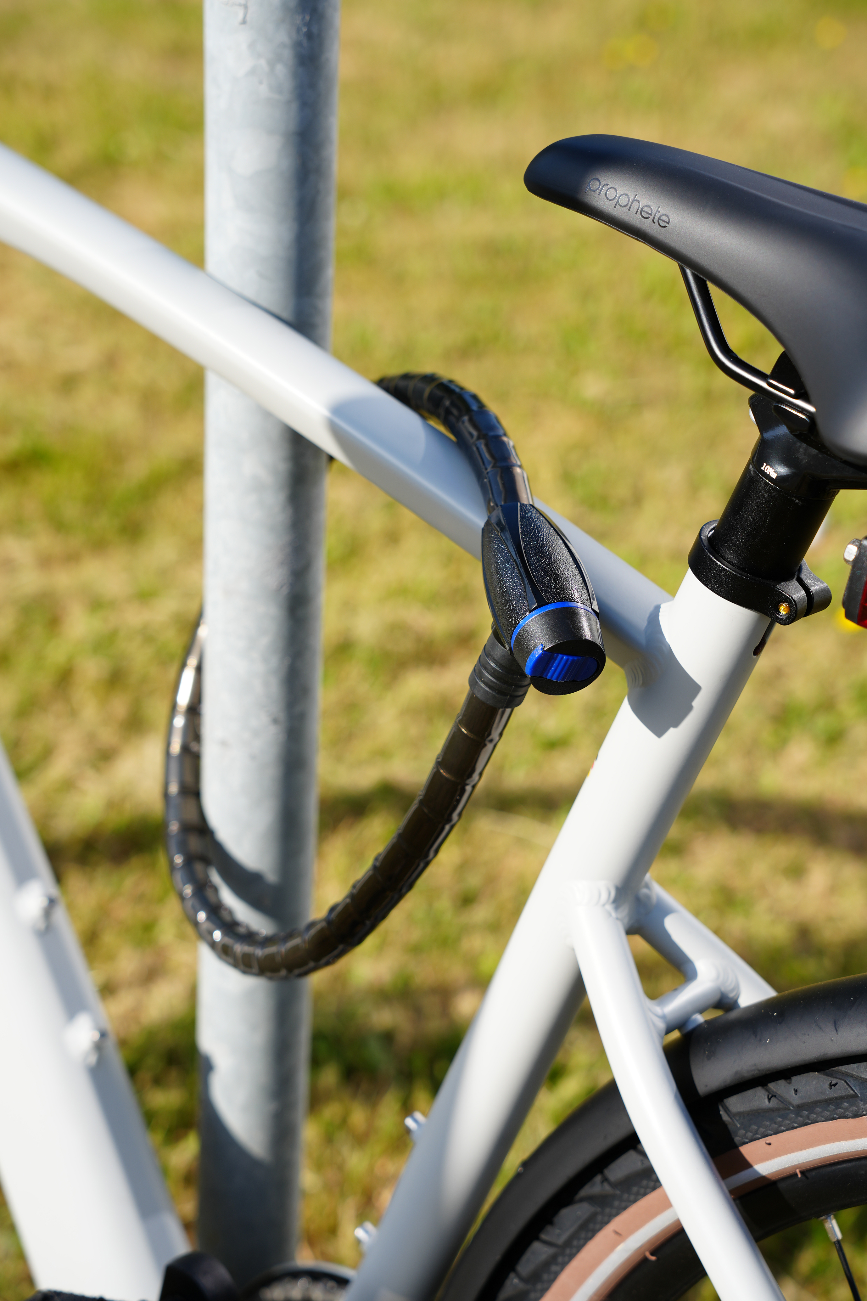 Ein Fahrrad, das mit einem schwarzen Schloss an einer Stange befestigt ist. Das Fahrrad ist weiß. Der Hintergrund ist Gras und blauer Himmel.