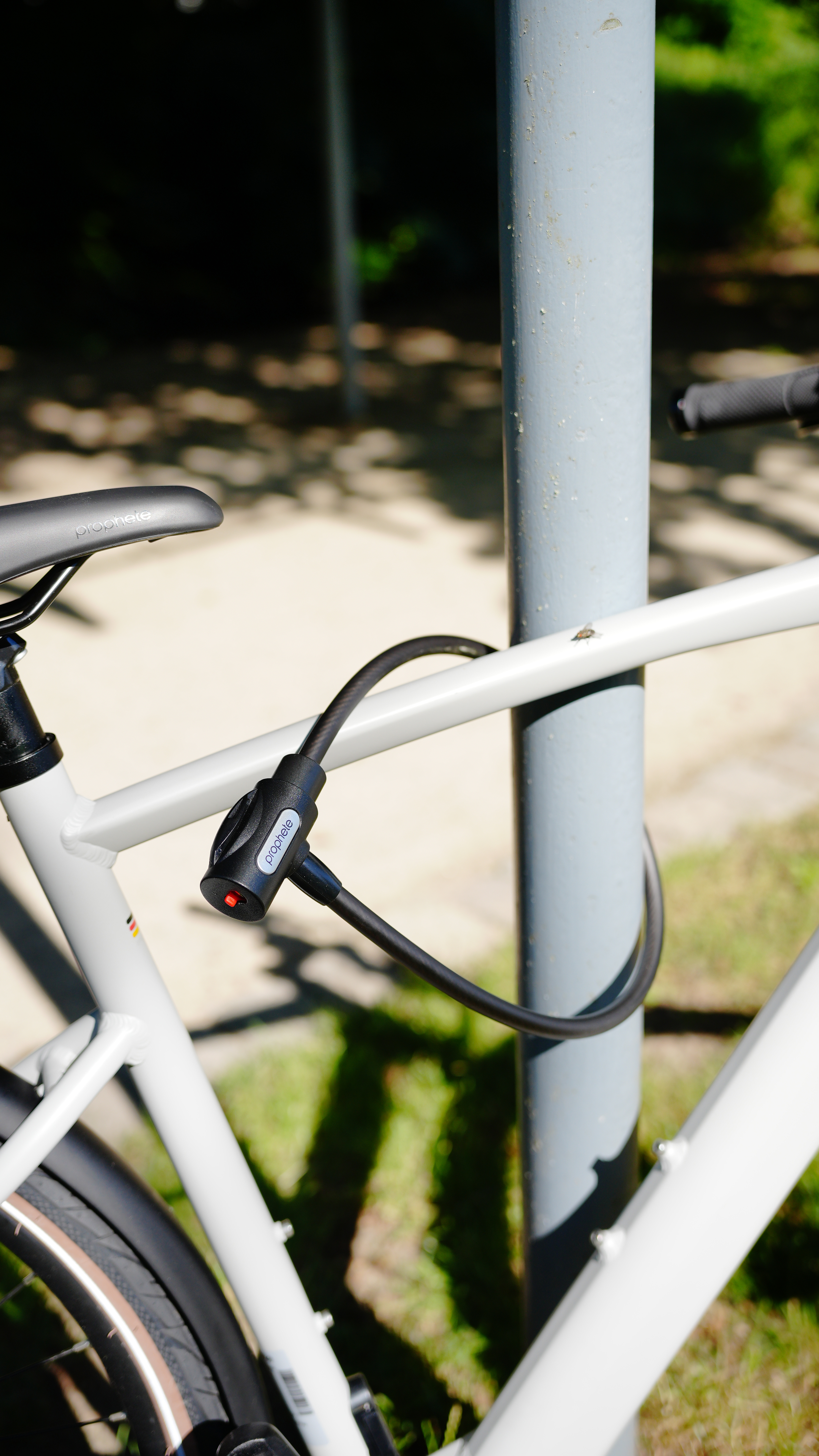 Ein Fahrrad, das mit einem schwarzen Kabelschloss an einem Metallpfosten gesichert ist. Die Szene spielt im Freien.