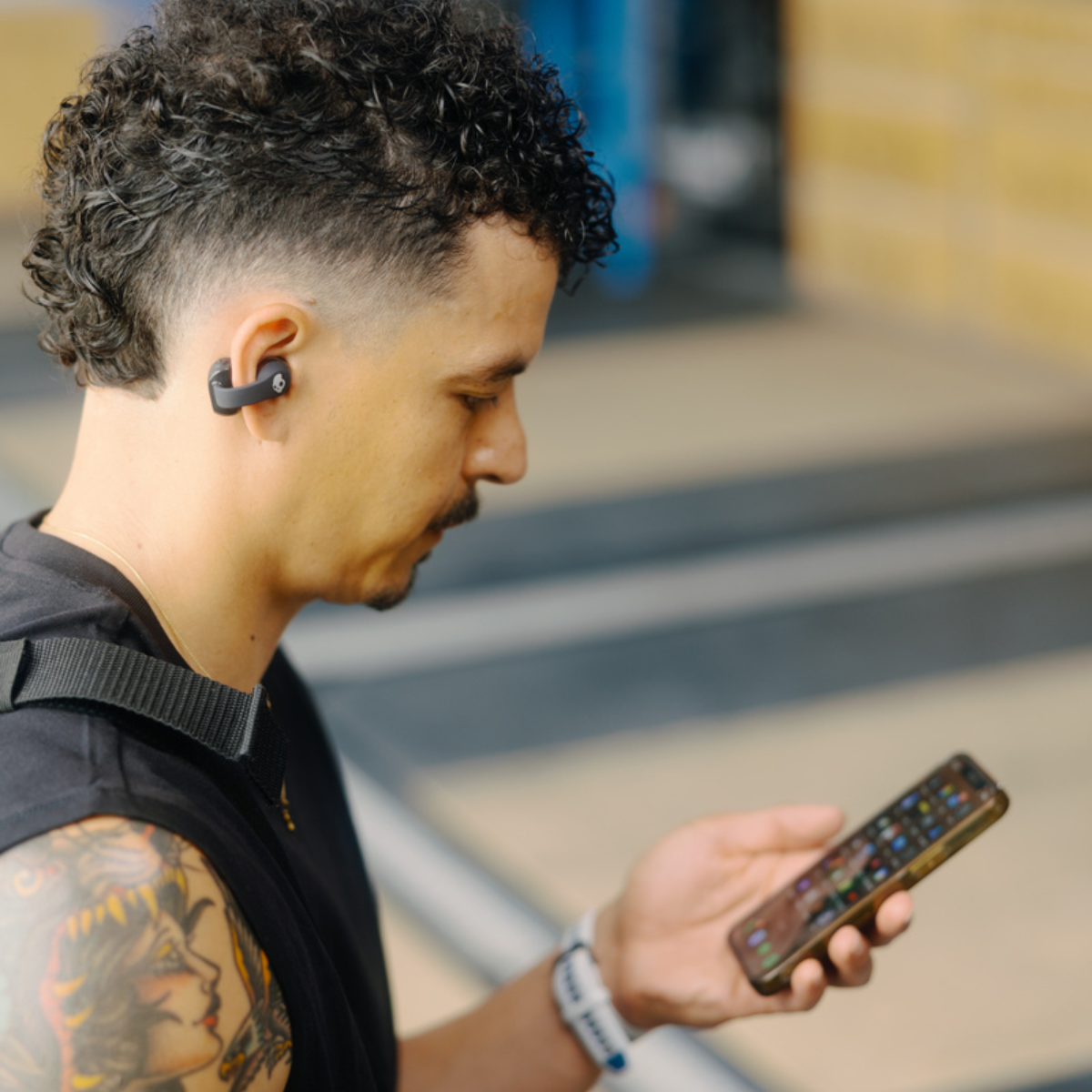 Un uomo con un telefono e auricolari. Ha tatuaggi e barba. Indossa una camicia nera.
