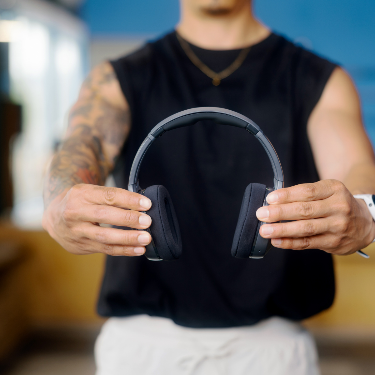 Osoba trzyma czarne słuchawki w stronę widza. Ma na sobie czarną koszulę i białe szorty. Rozmyte tło.