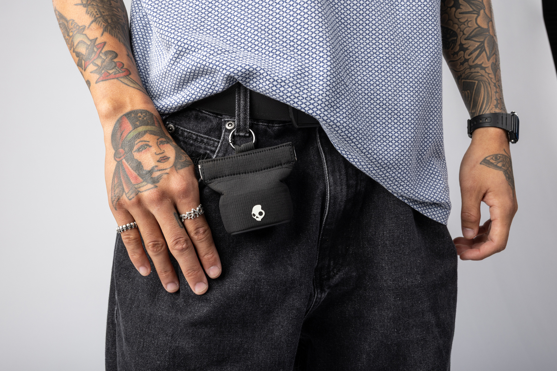 Mano tatuata tiene una custodia nera attaccata a una cintura. Indossano jeans scuri. Sfondo grigio.
