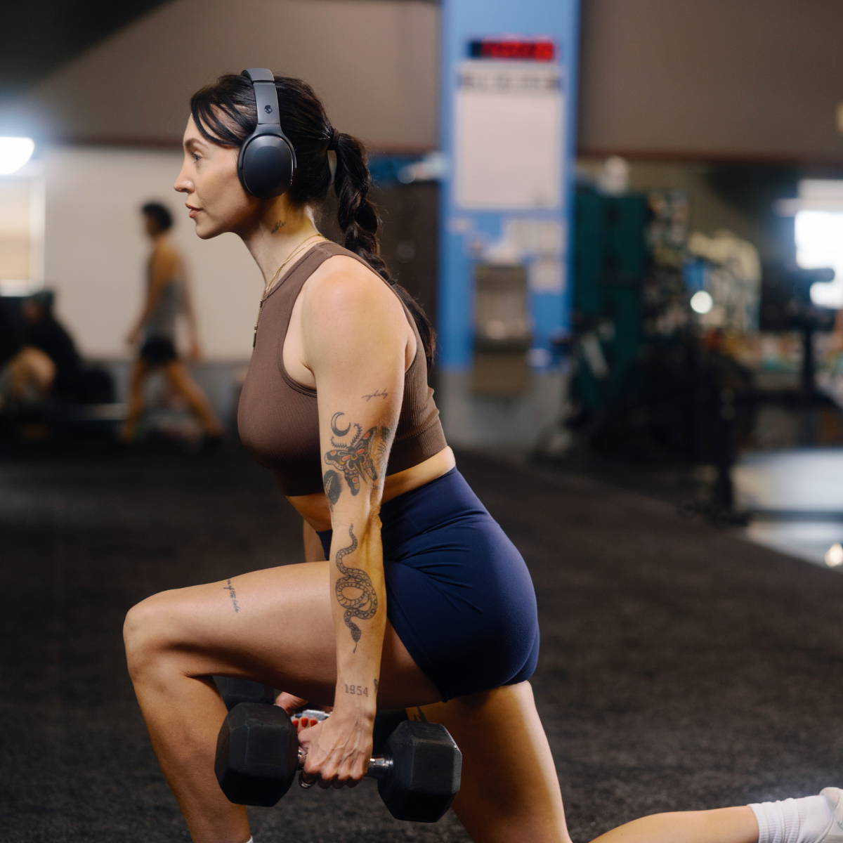 Kobieta robi wykroki w czarnych słuchawkach na siłowni, trzymając hantle.